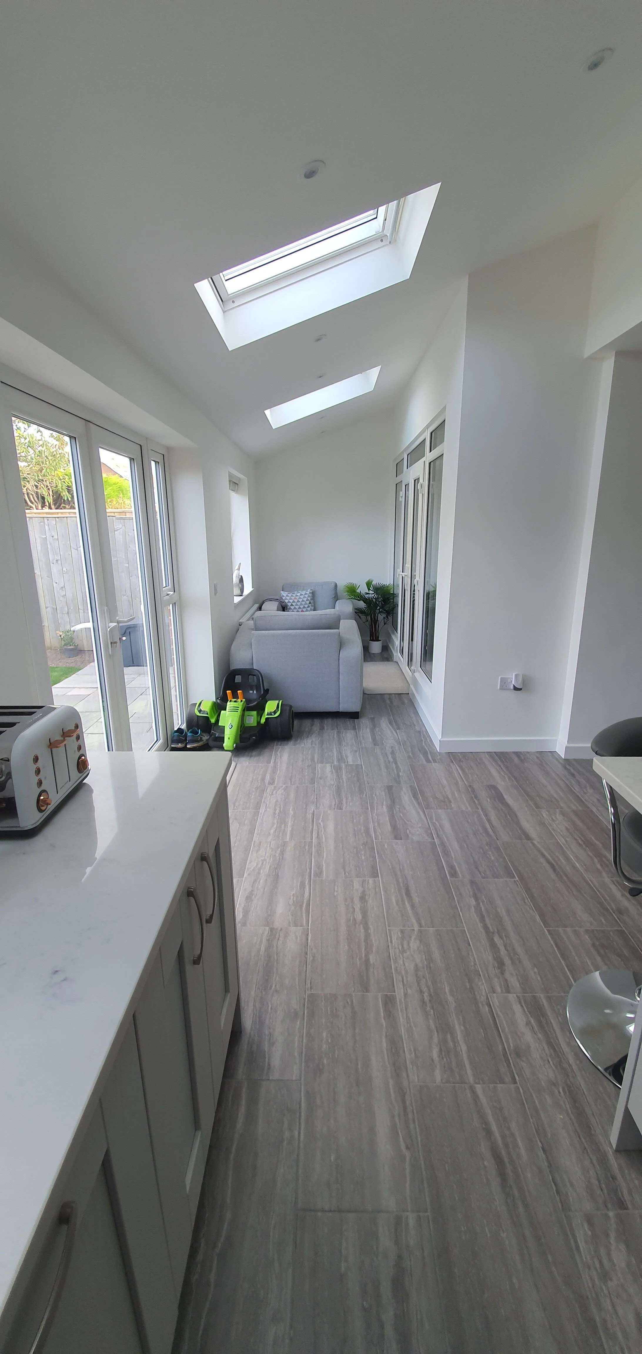 A bright, modern sunlit living area with a gray sofa, potted plant, and windows/doors leading outside. Skylights in the ceiling provide additional natural light. Children's toys are nearby.