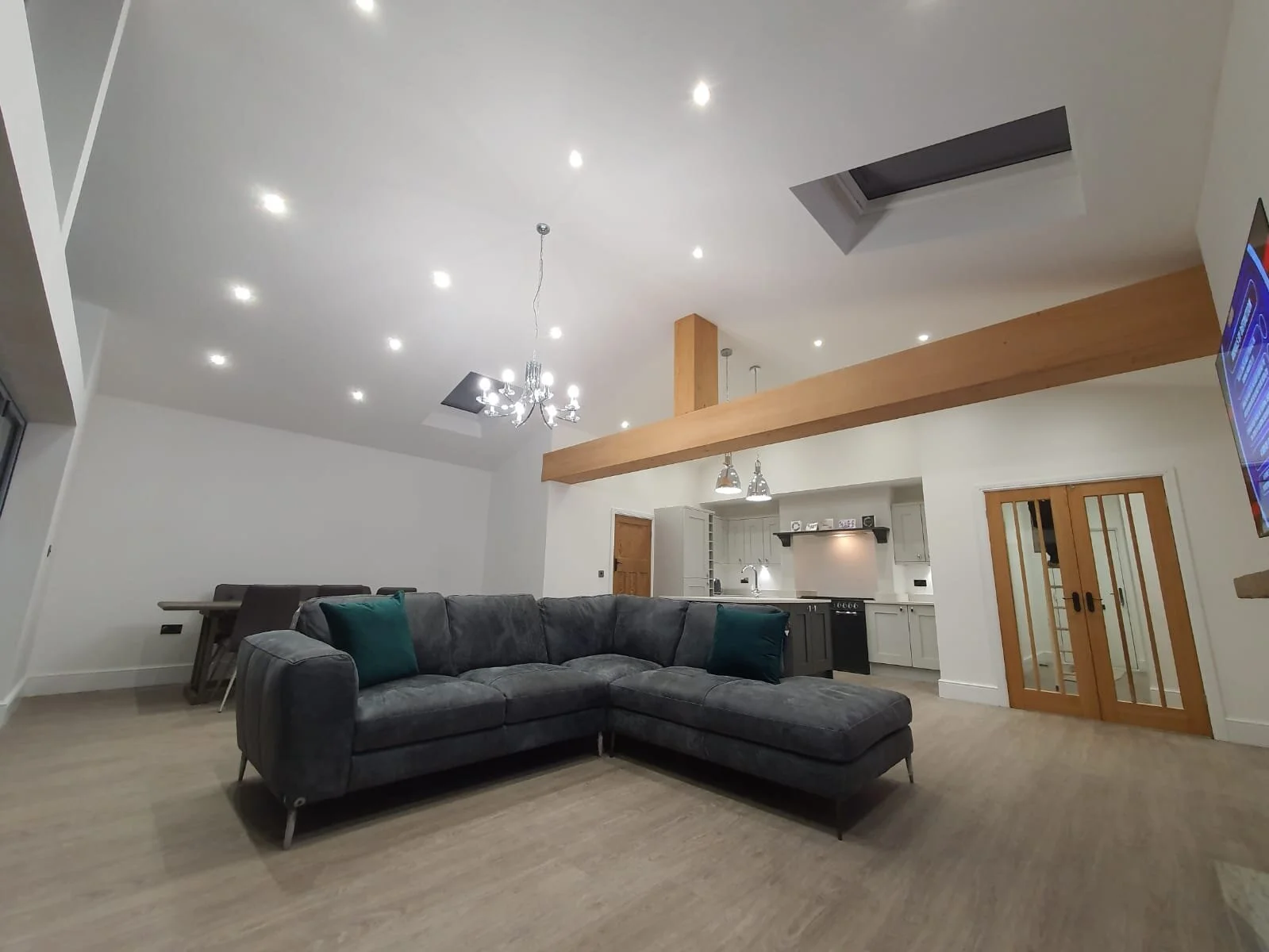 Living room with gray sectional sofa, teal cushions, wooden flooring, white walls, open kitchen with white cabinets, black appliances, two pendant lights, wooden beam, glass sliding doors, and a wall-mounted TV.
