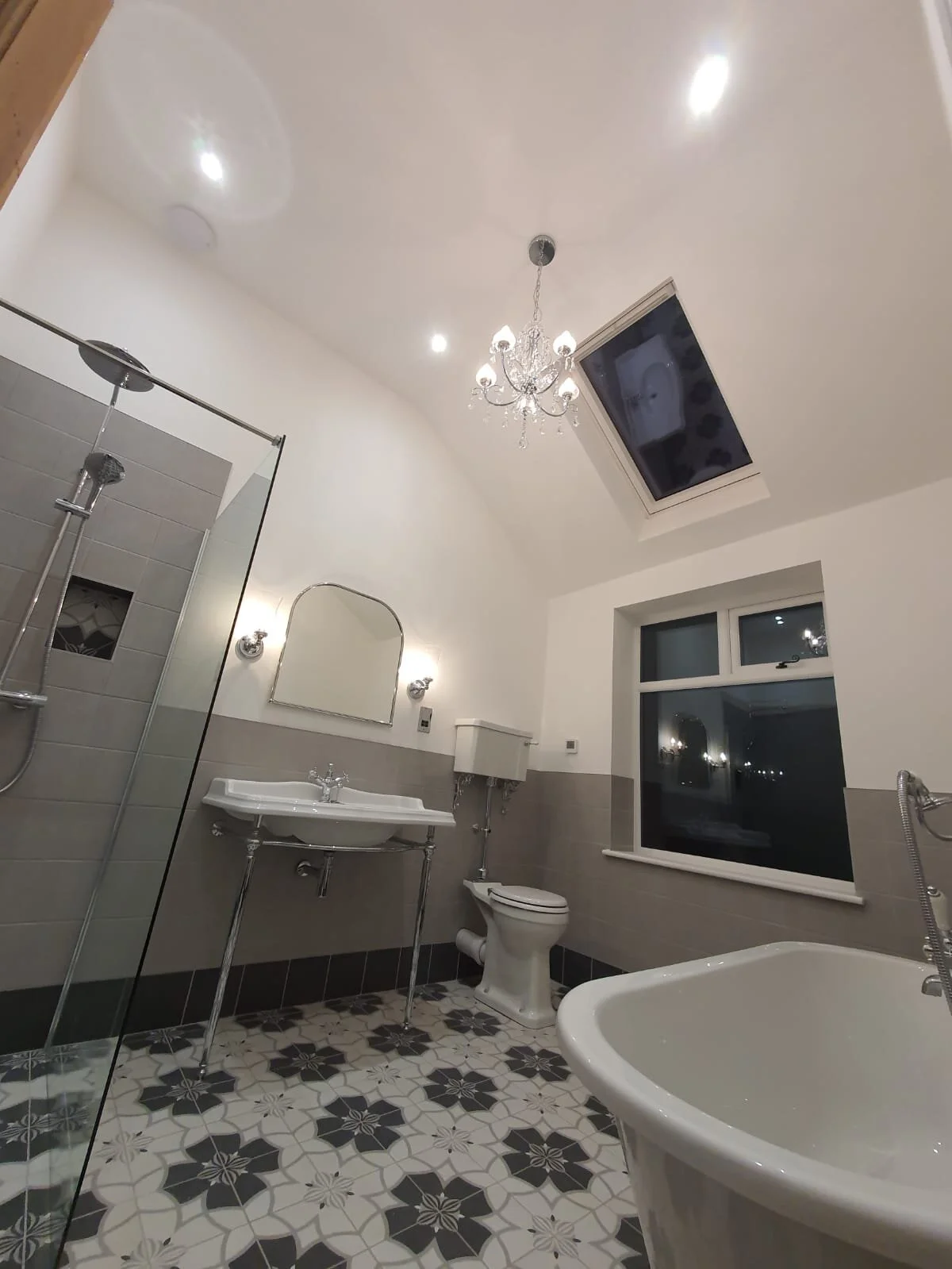 A bathroom featuring a shower with glass doors, a sink with a mirror, a toilet, a bathtub, a skylight, a window, and a chandelier on the ceiling with white walls and decorative tiled floor.