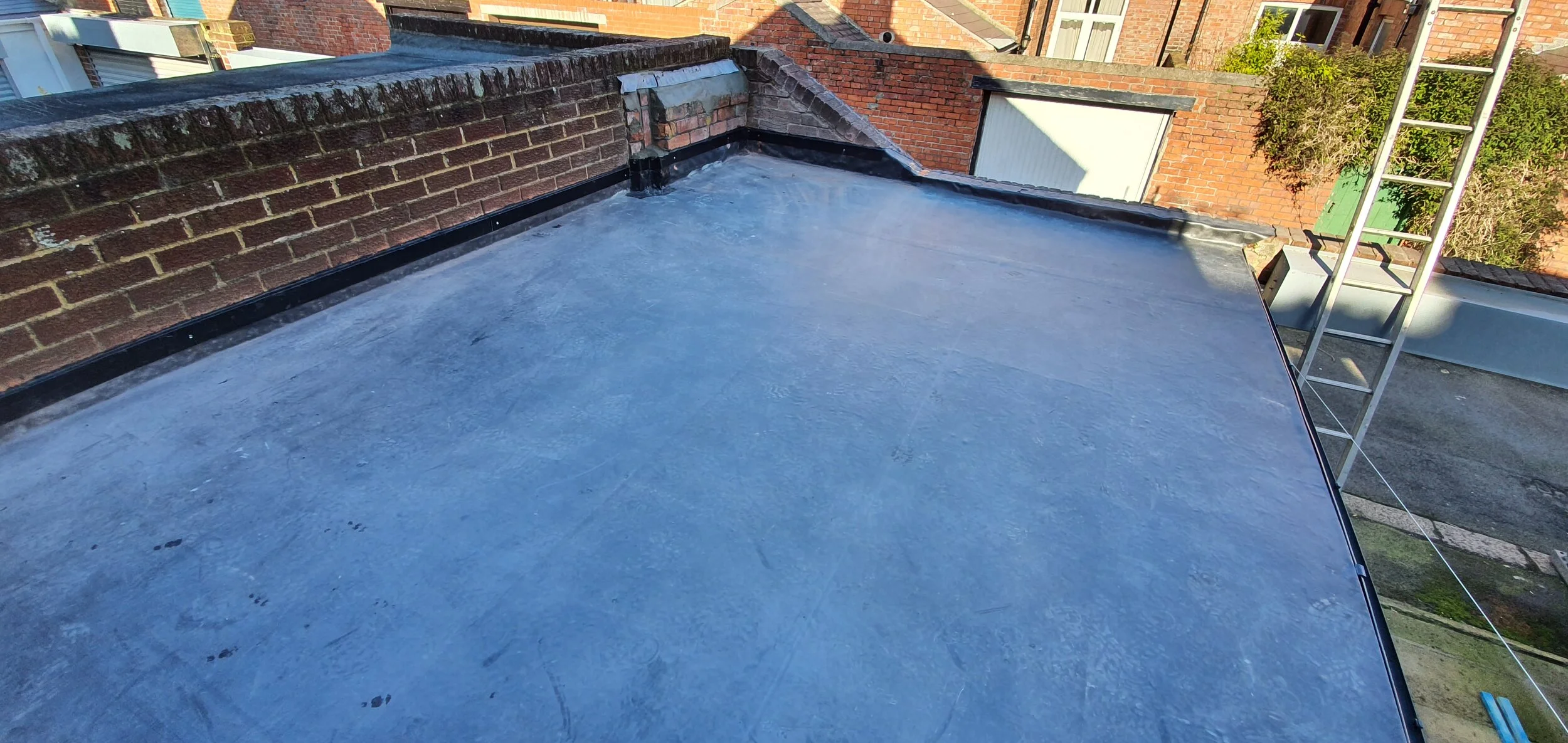 Rooftop with flat waterproof surface, brick wall, ladder, and neighboring buildings and trees.