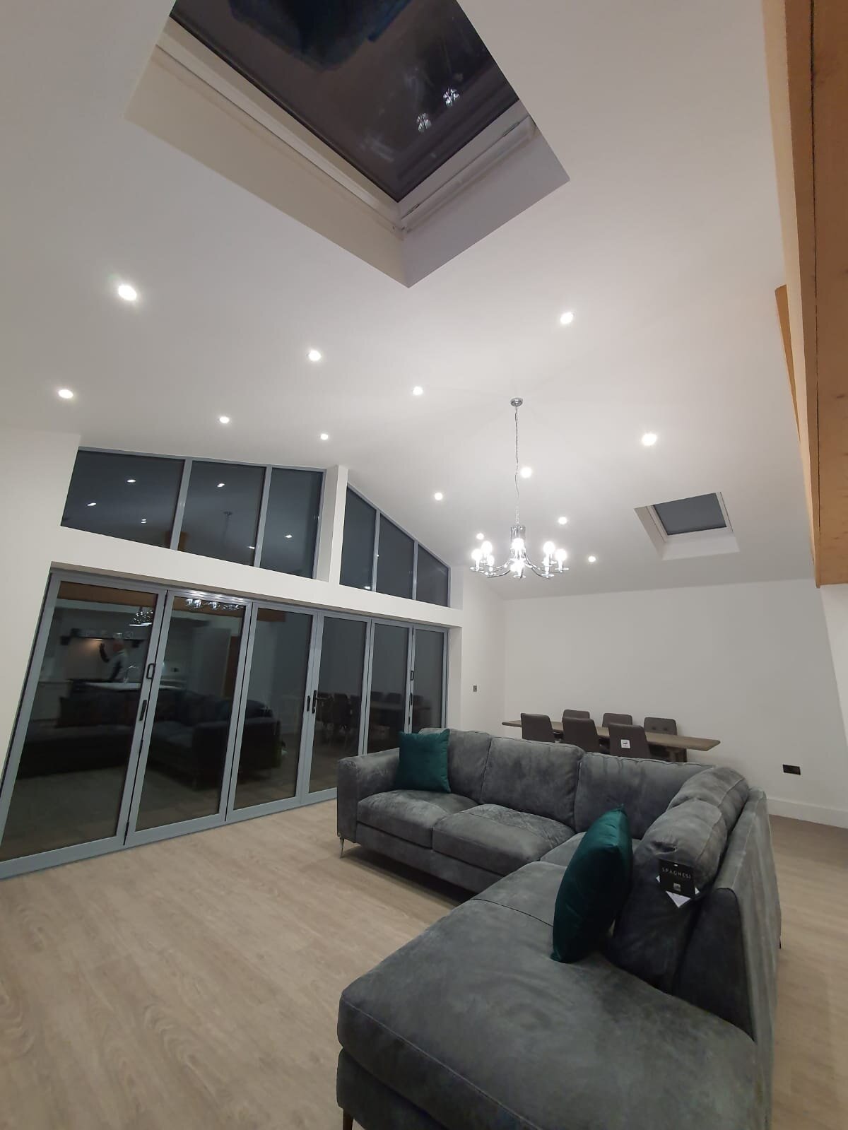 Modern living room with a grey sectional sofa, a wooden dining table with chairs, sliding glass doors, high ceiling with a chandelier, and two skylights in the ceiling.