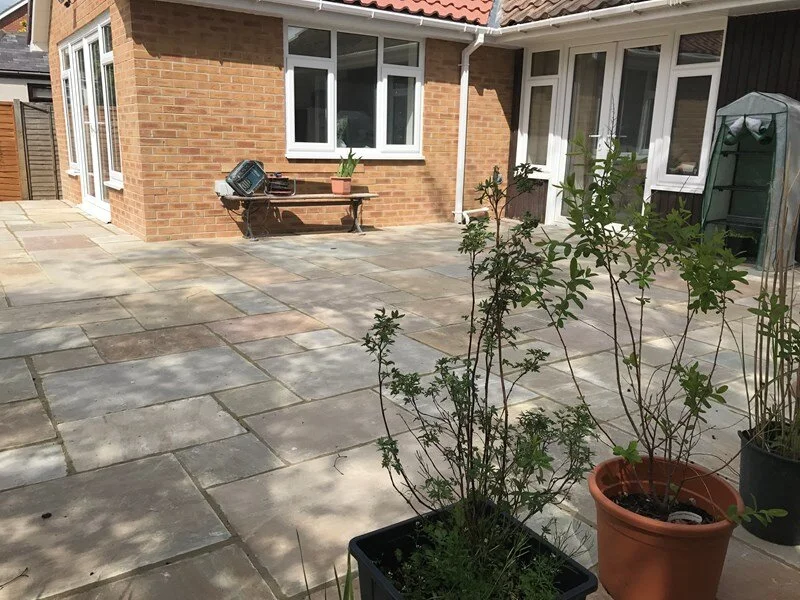 A backyard patio with potted plants, a brick house with white window frames, a wooden bench with a potted plant and a grill on it, and a greenhouse in the background.