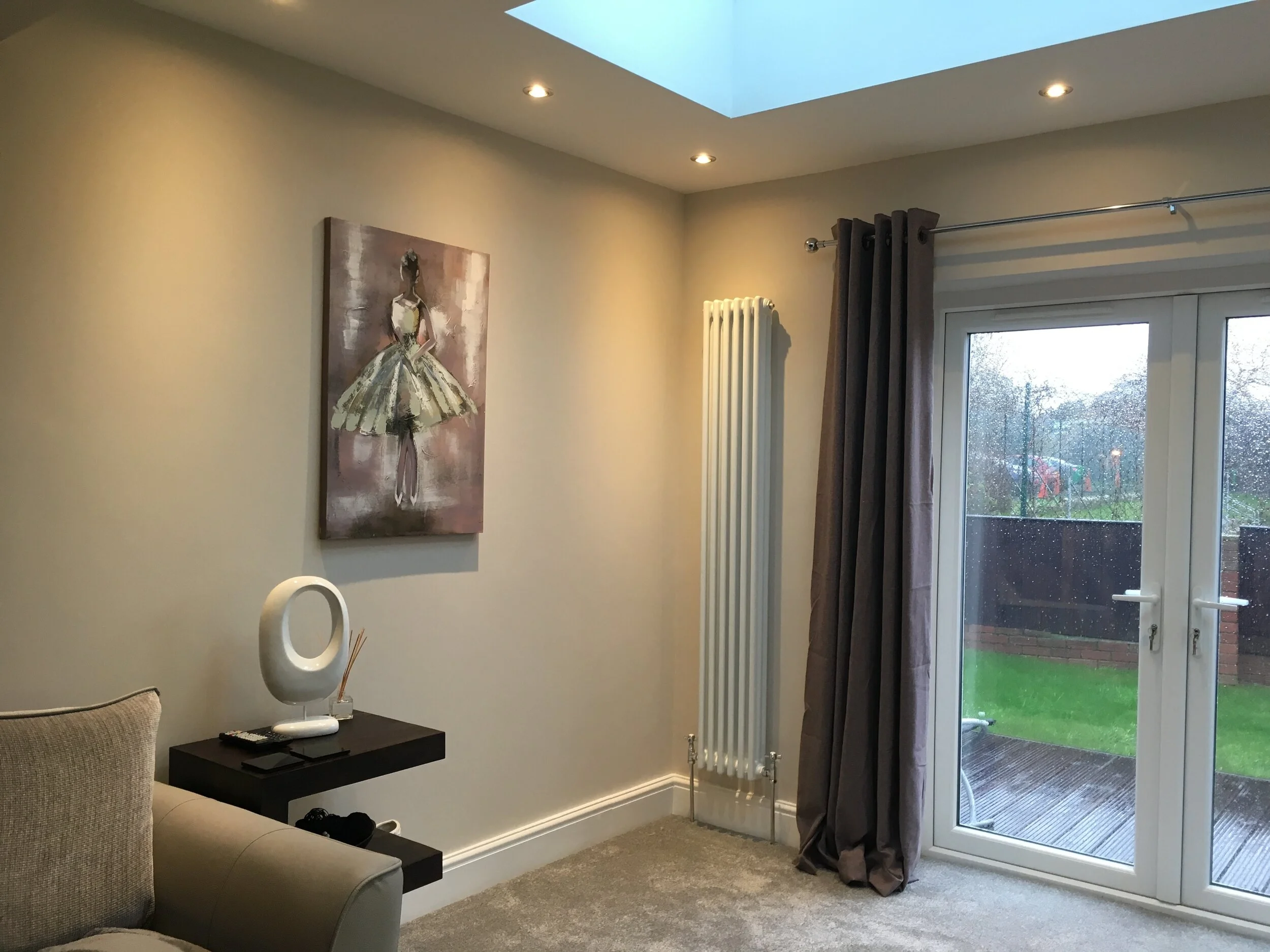 Living room with beige walls, a painting of a ballerina dress, a black shelf with a white modern decorative lamp, and glass doors with brown curtains leading to a patio, on a rainy day.