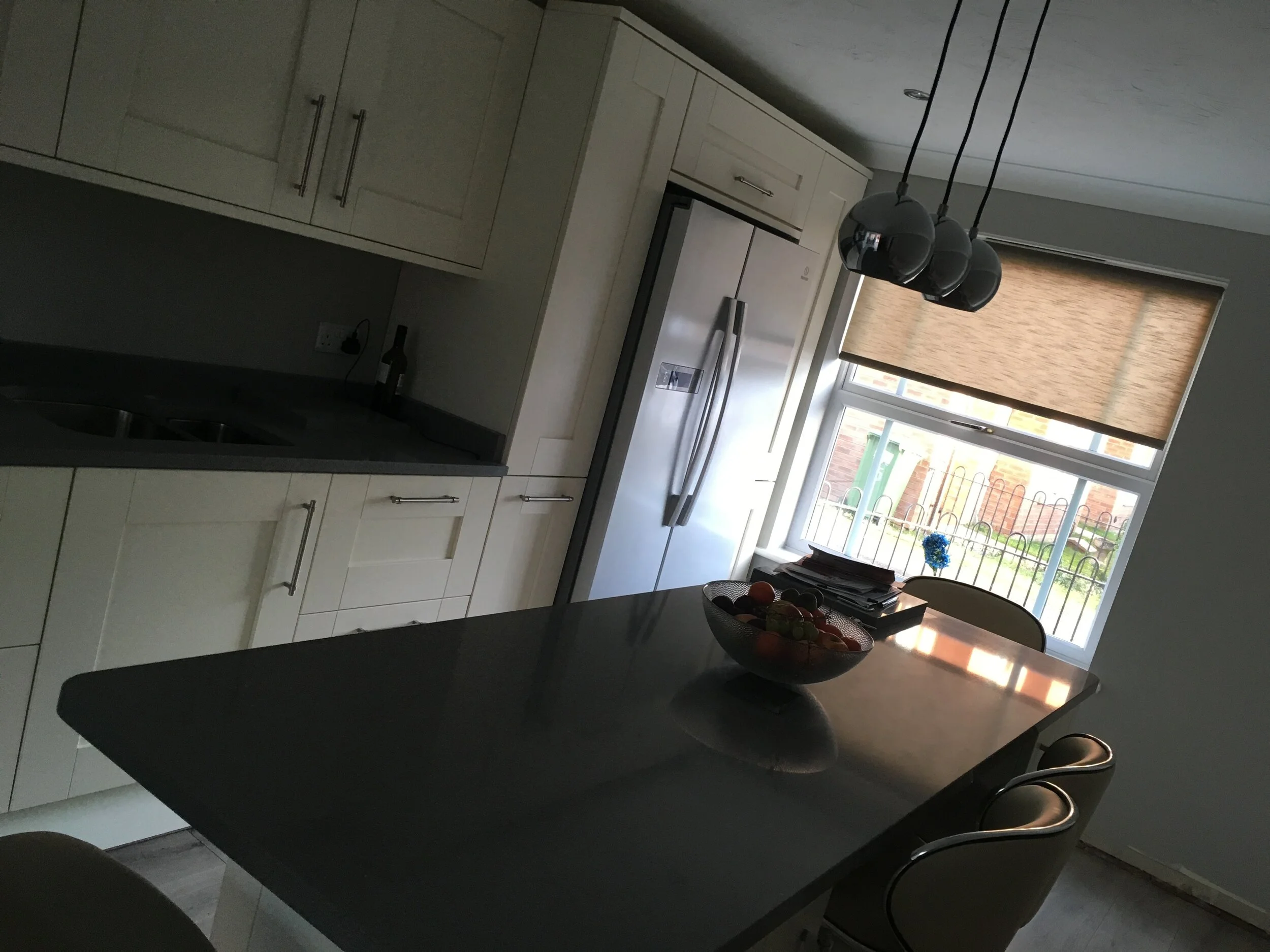 A kitchen with a dark countertop, light cabinets, a stainless steel refrigerator, a window with a brown blind, a bowl of fruit, and hanging pendant lights.