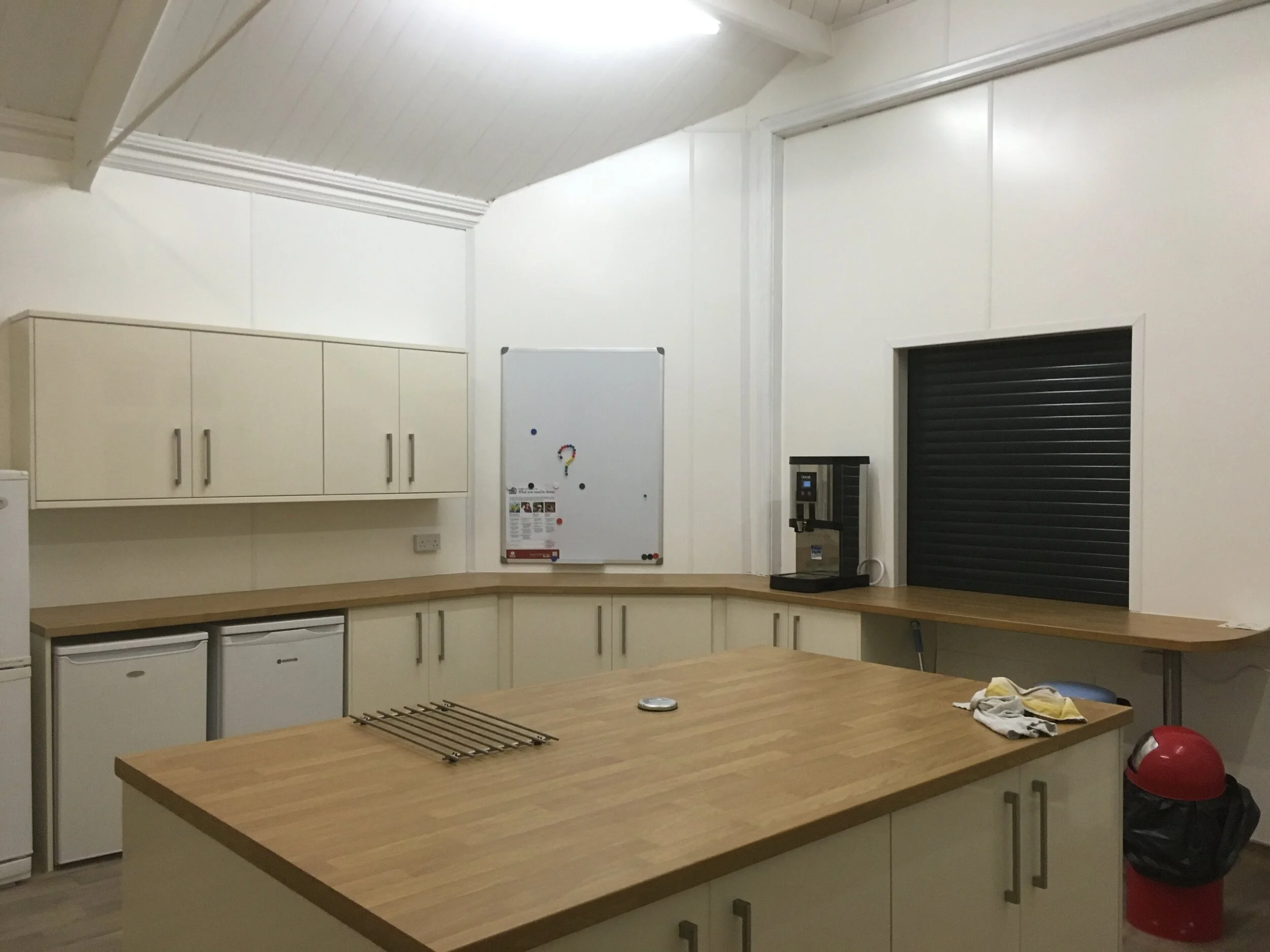 Empty commercial kitchen with wooden countertops, white cabinets, a small fridge, a coffee machine, and a whiteboard on the wall.