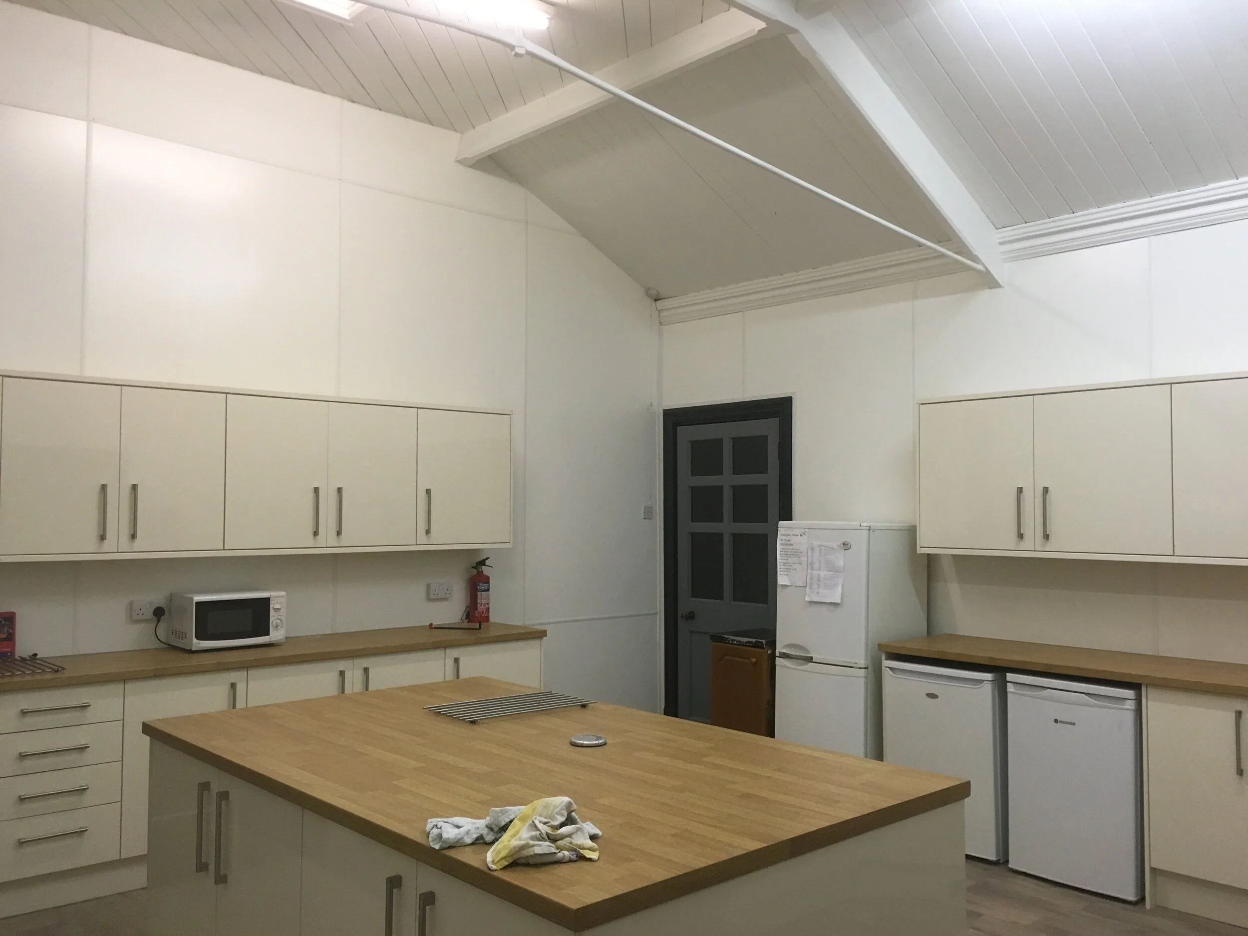 A kitchen with white cabinets, a wooden countertop on a kitchen island, a microwave, a fire extinguisher, a refrigerator, and small white fridges, under a sloped ceiling.