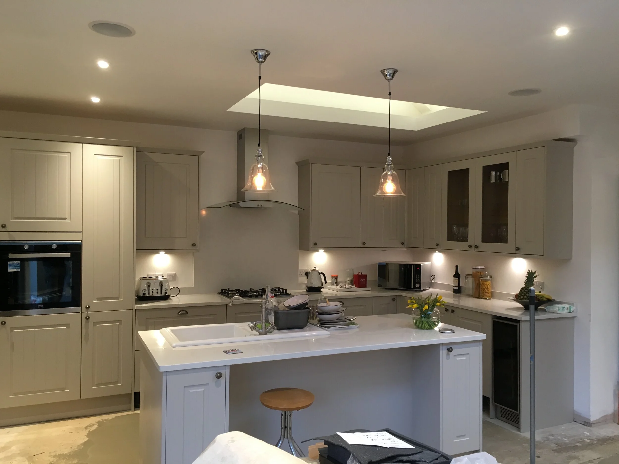 Modern kitchen with beige cabinets, white countertops, two hanging pendant lights over a central island, built-in oven, microwave, gas cooktop, and various small appliances. Decorative plants and fruit on the counter, and a stool at the island.