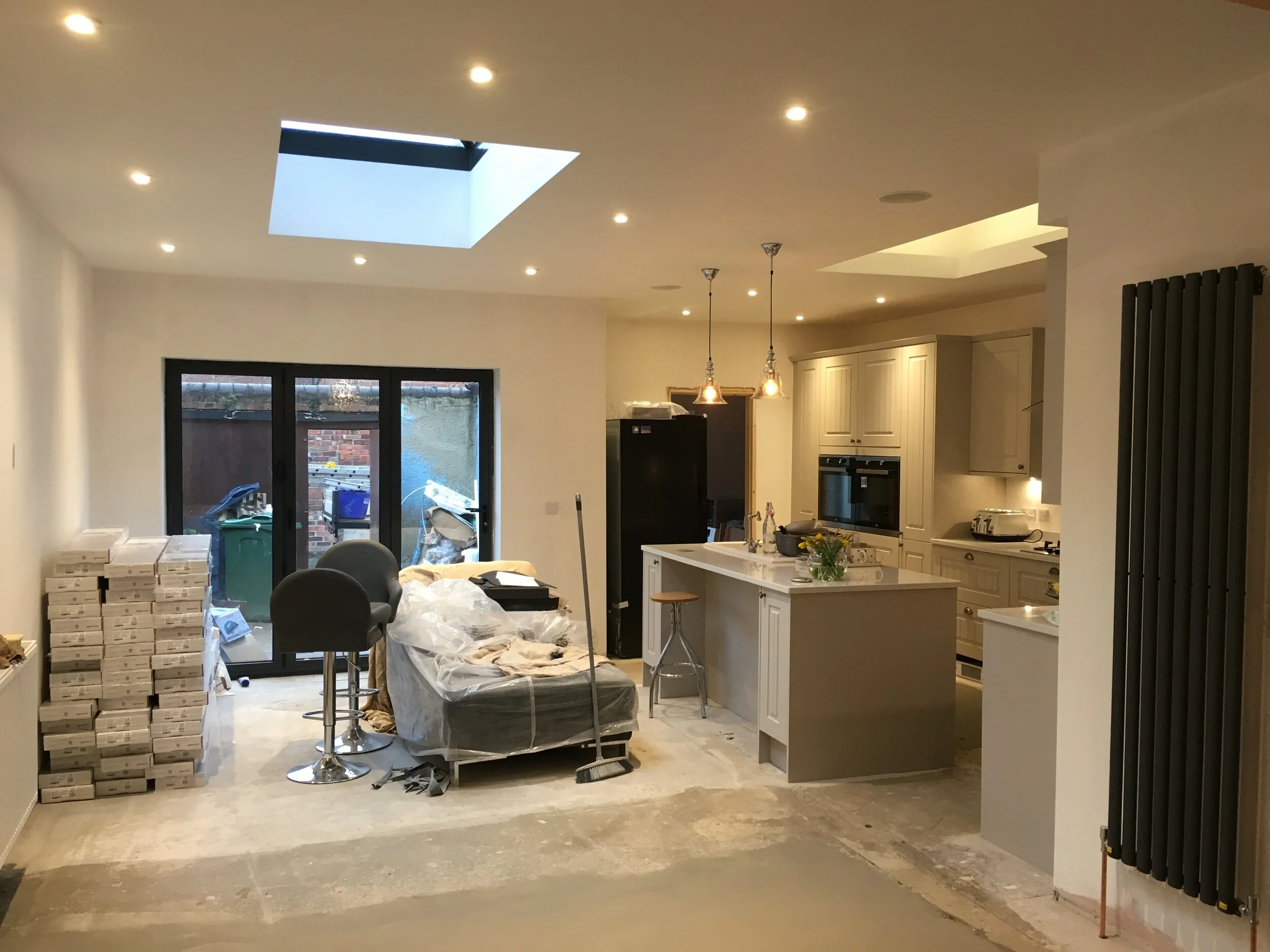Interior view of a modern kitchen and living room under renovation, with stacked boxes, plastic-covered furniture, and construction supplies.