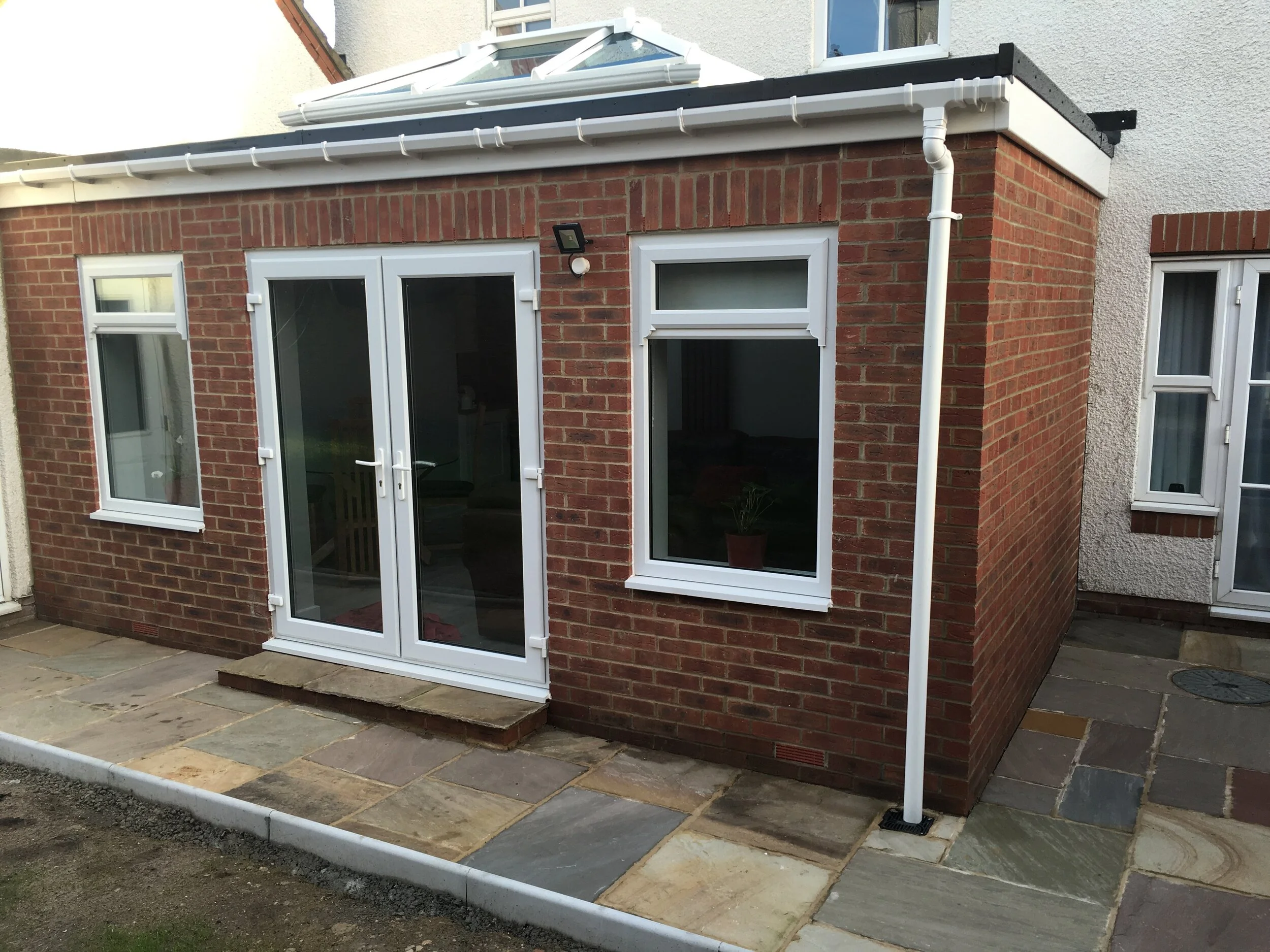 Back of a house with red brick walls, a white glass door, and two white-framed windows. There is a small stone step leading to the door, and a stone patio in front. A gutter pipe goes from the roof to the ground.