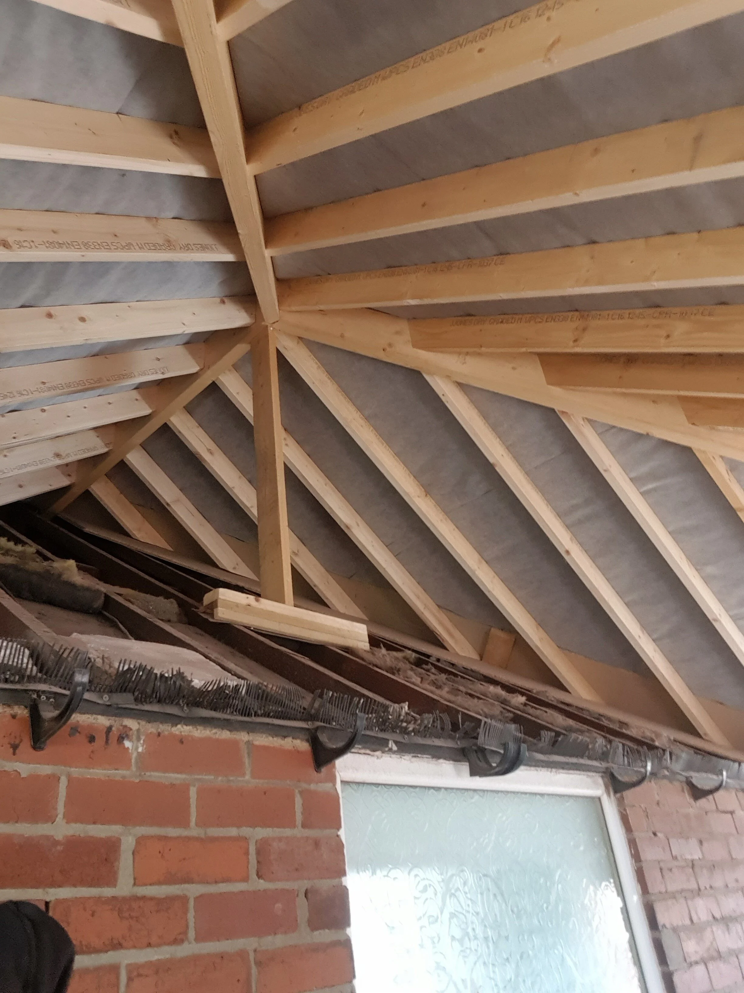 Interior of a building under construction, showing roof framing with wooden trusses and insulation, brick wall, and a frosted window.