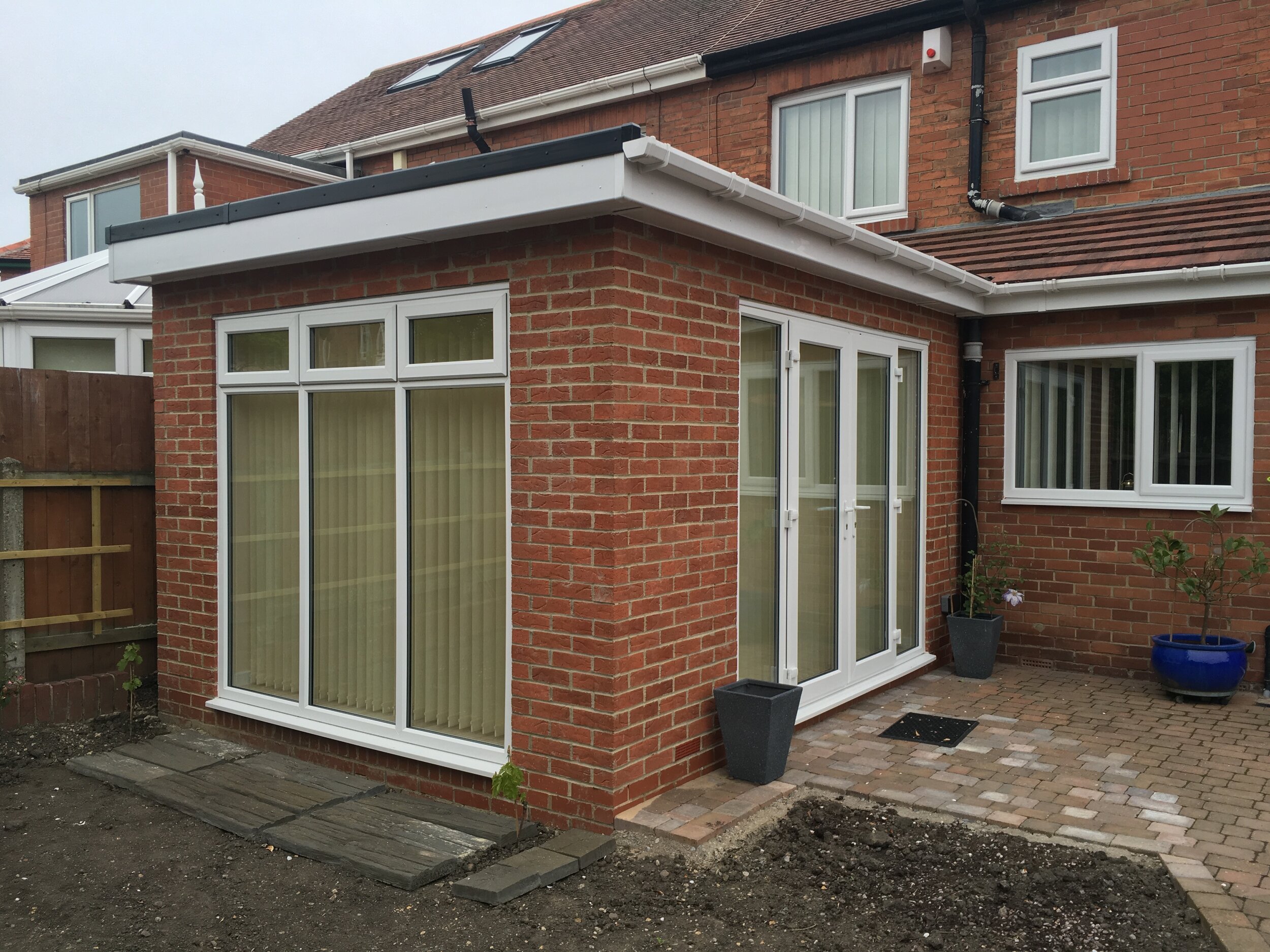 Backyard view of a brick house with large windows and sliding glass doors, some potted plants, a brick patio, and a garden area with dirt and paving stones.