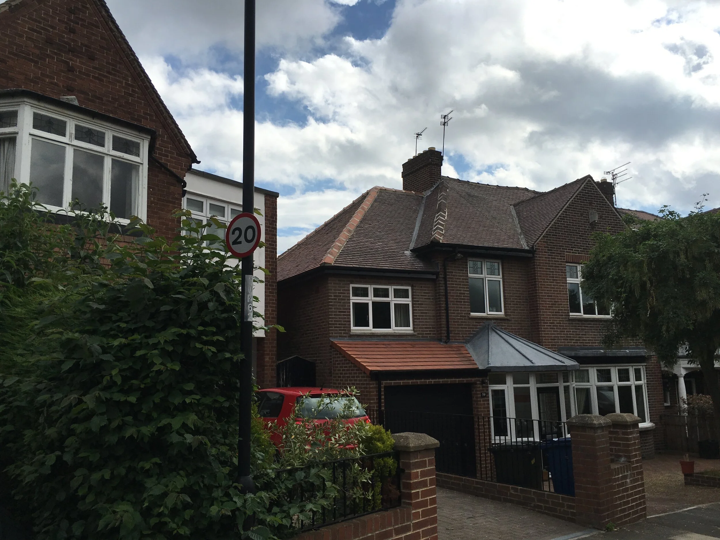 Two-story brick house with bay windows, a garage with a red-tiled roof, a car parked in front, and a 20 mph speed limit sign on a street corner under a cloudy sky.