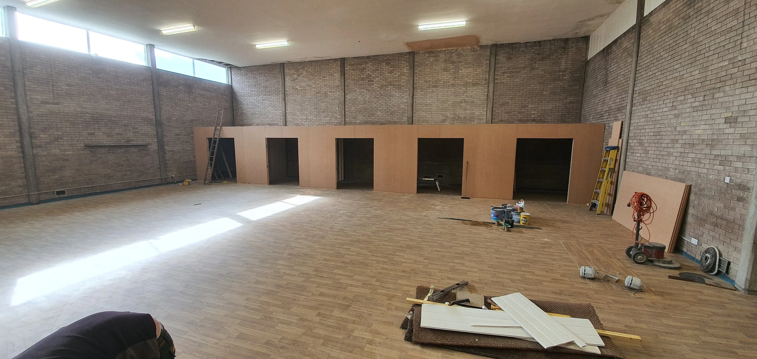 Interior of a large room under construction with unfinished brick walls, a wooden partition with four open doorways, construction tools, materials, and equipment scattered on the floor.