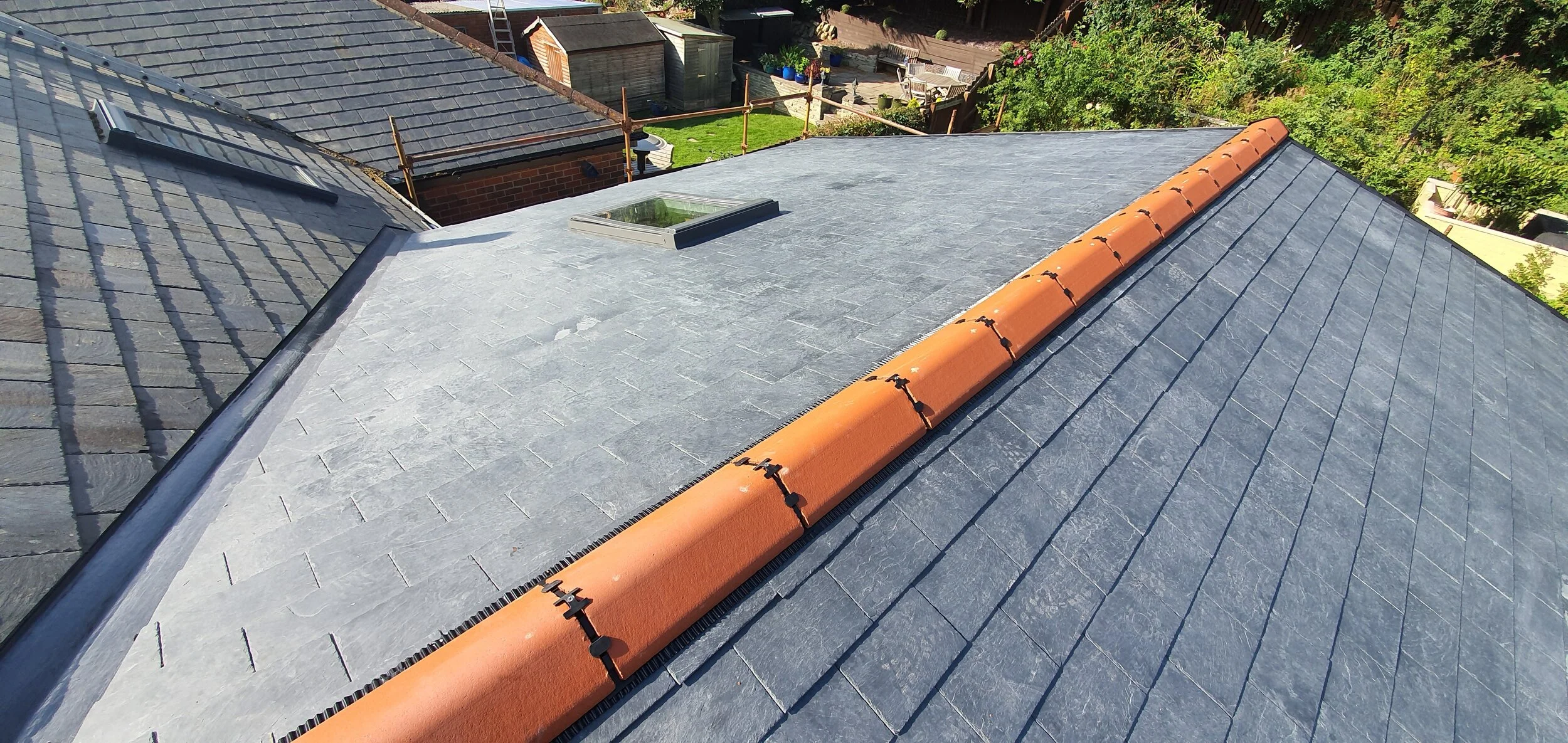 Aerial view of a house roof with slate tiles, a central ventilation skylight, and a ridge with orange tiles, surrounded by neighboring rooftops and greenery.