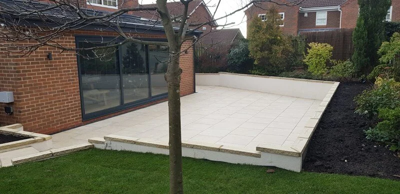 Backyard patio with white tiles, a tree, a brick house with sliding glass door, and surrounding greenery.
