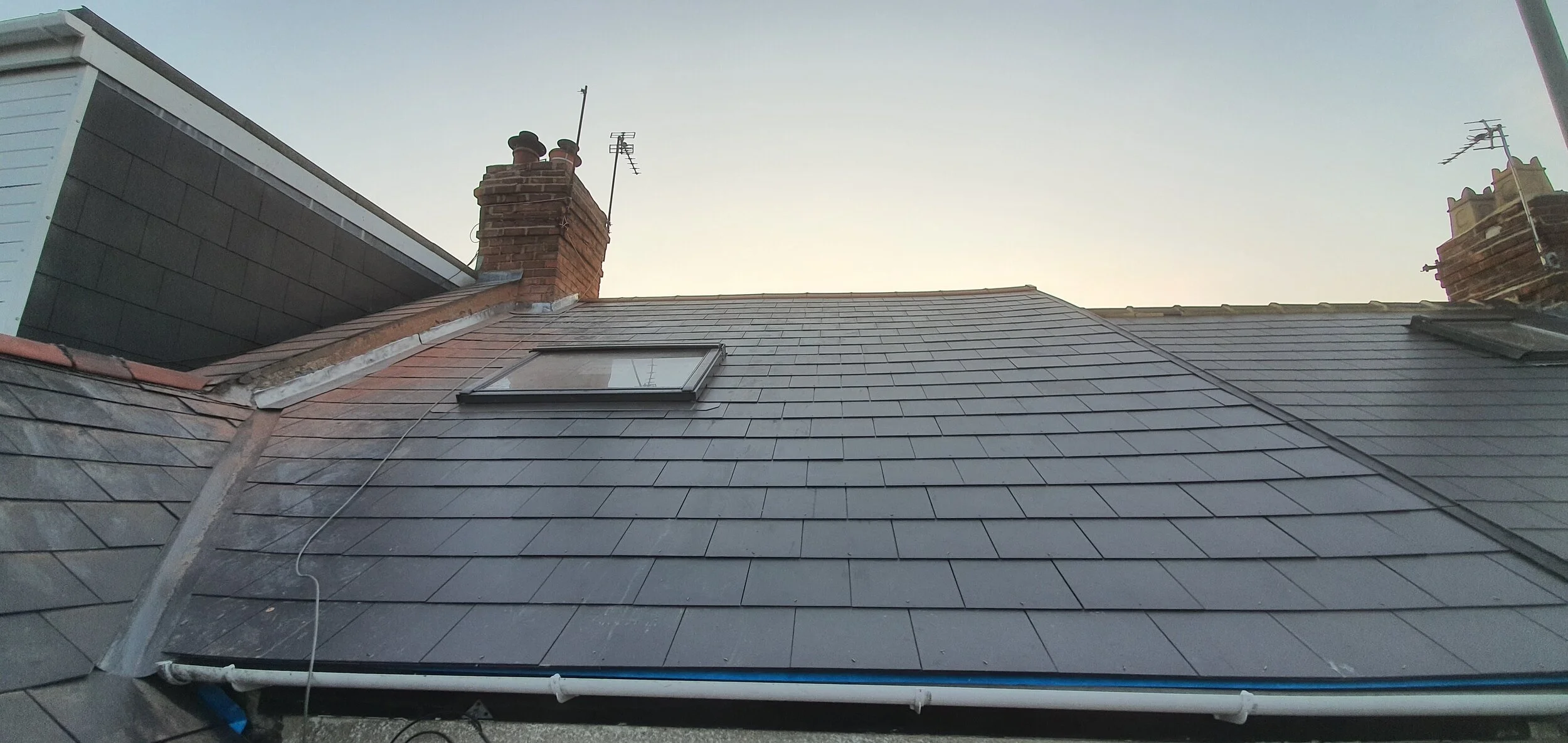 Photo of a rooftops and chimneys, including a skylight window on the slate roof, with a clear sky in the background.