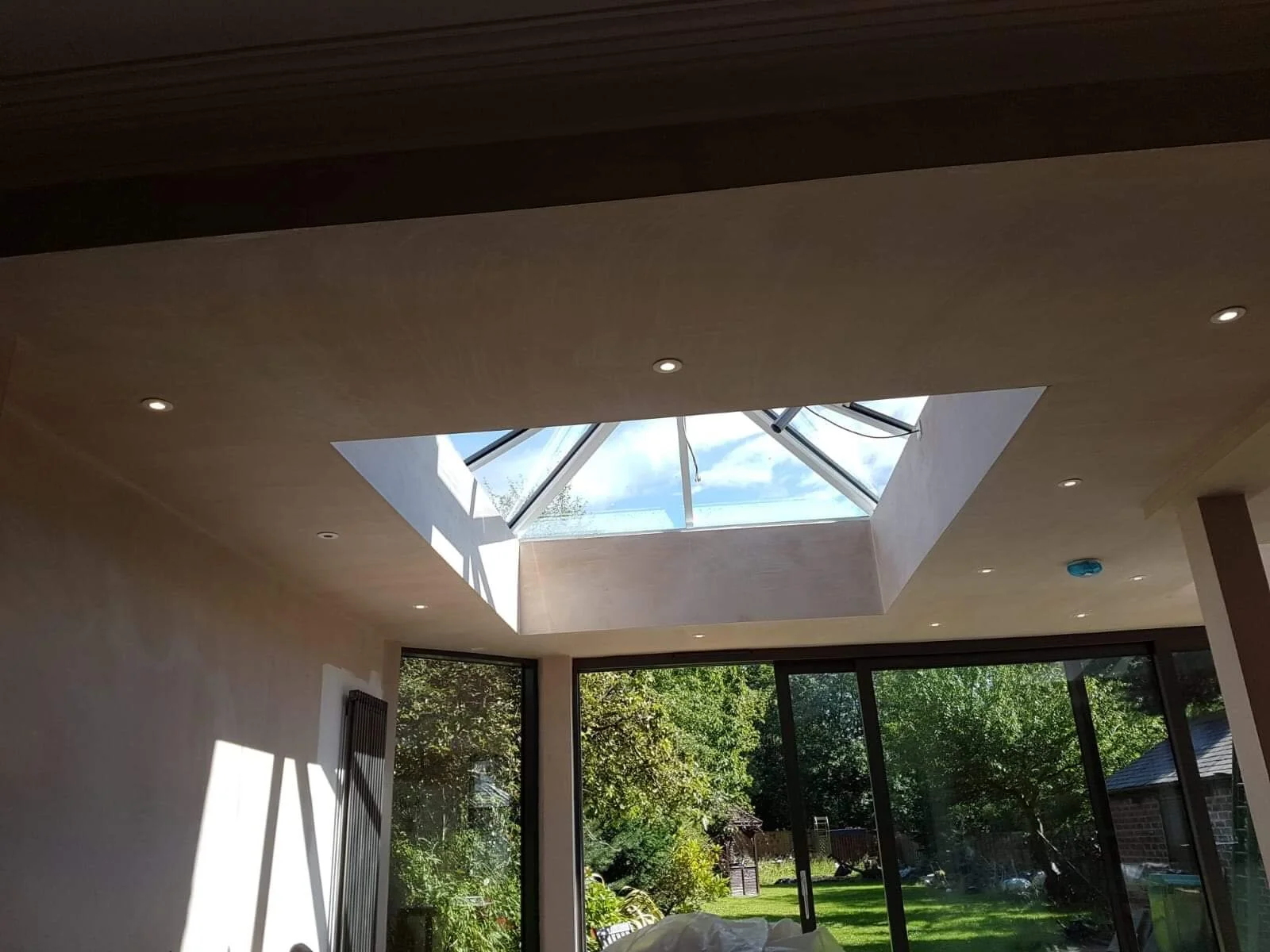 Interior view of a room with large glass windows and a skylight, showing a garden outside with trees and a lawn.