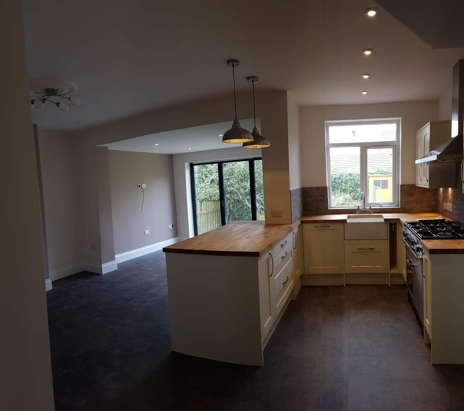 Empty modern kitchen with white cabinets, wooden countertops, a window above the sink, and a brick backsplash, with a dining area and sliding glass door leading outside.