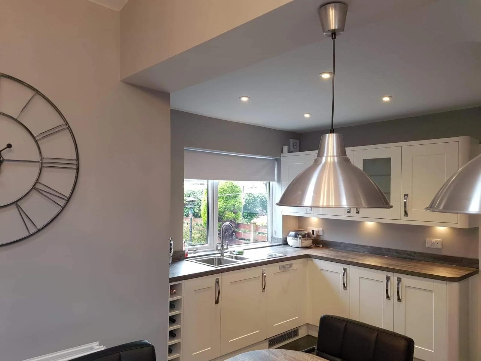 Modern kitchen with white cabinets, dark countertop, stainless steel pendant lights, a window above the sink, and a wall clock on the left.