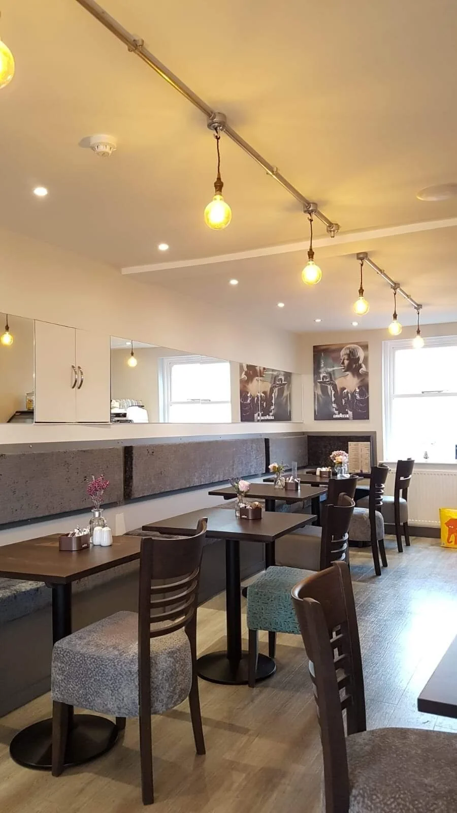 Interior of a cozy cafe or restaurant with wooden tables, chairs, floral centerpieces, wall art, and warm lighting.