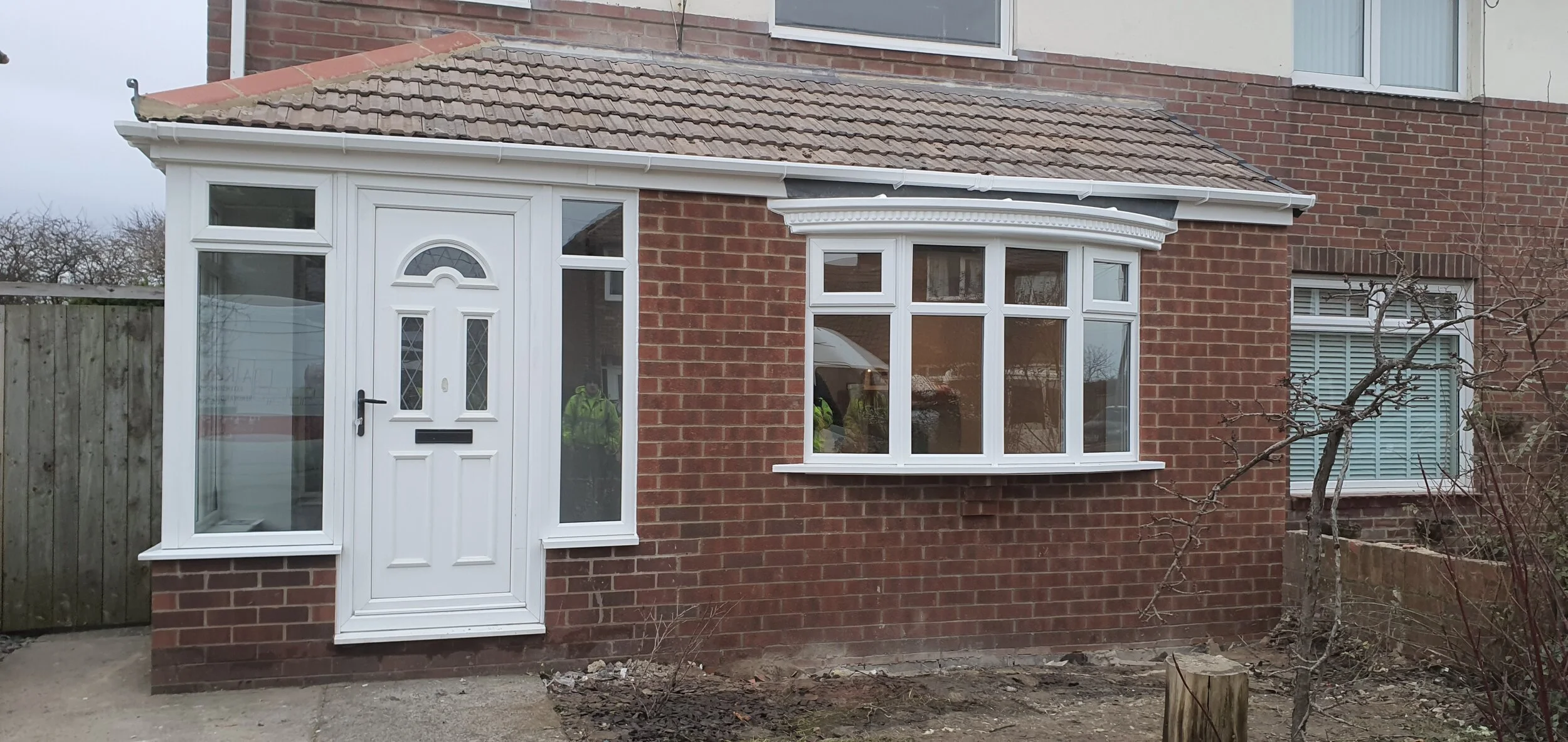 Front exterior of a brick house with a white enclosed porch, featuring a white door with window panels, a bay window, and a small leafless tree in the garden.