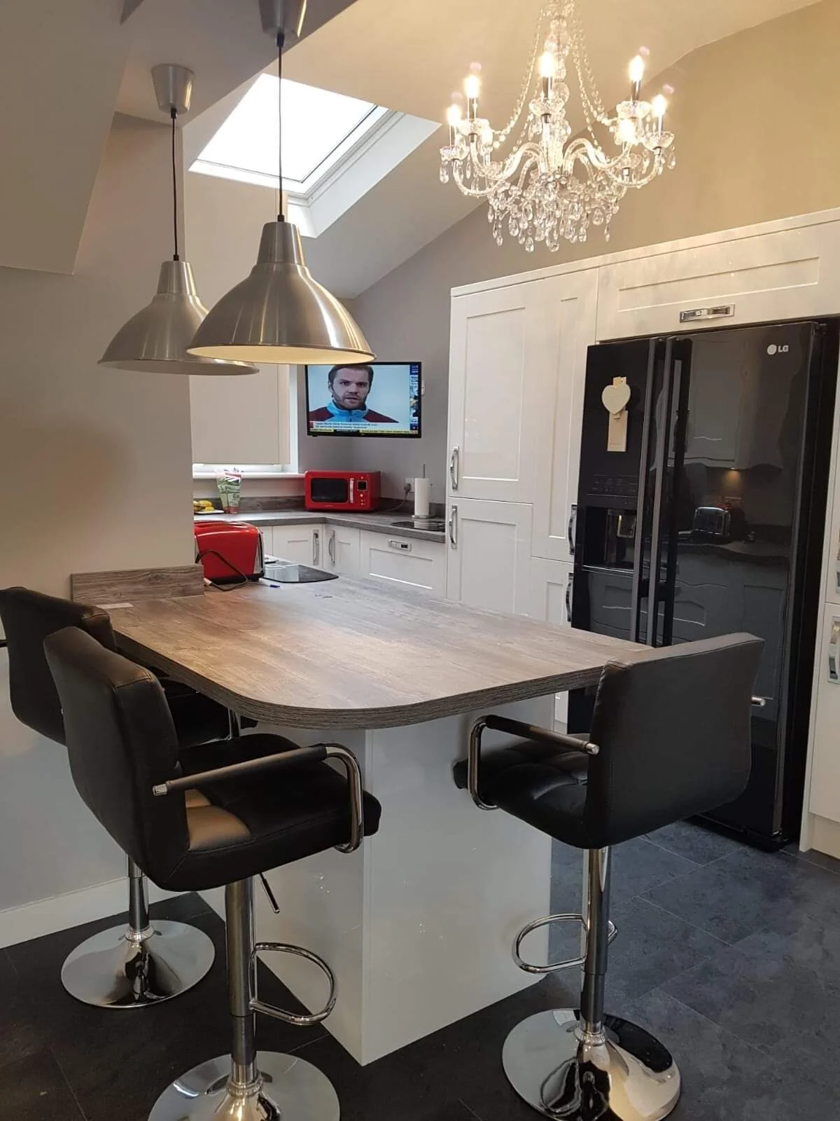 Modern kitchen with a wooden island, black leather bar stools, white cabinetry, black refrigerator, red toaster, microwave, and a hanging chandelier with three pendant lights, under a skylight and a flat-screen TV on the wall.
