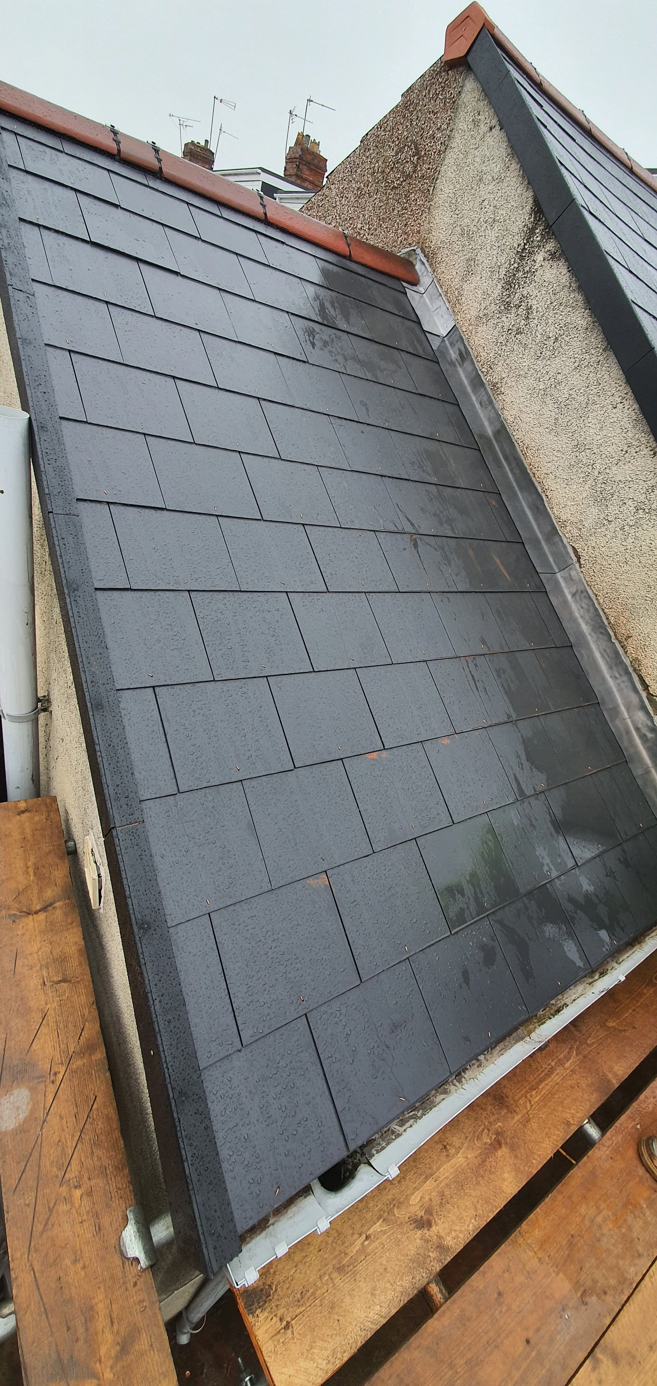 The image shows a rooftop with newly installed dark grey shingles, wet from rain, and adjacent to an older, weathered wall and roof with different roofing materials.