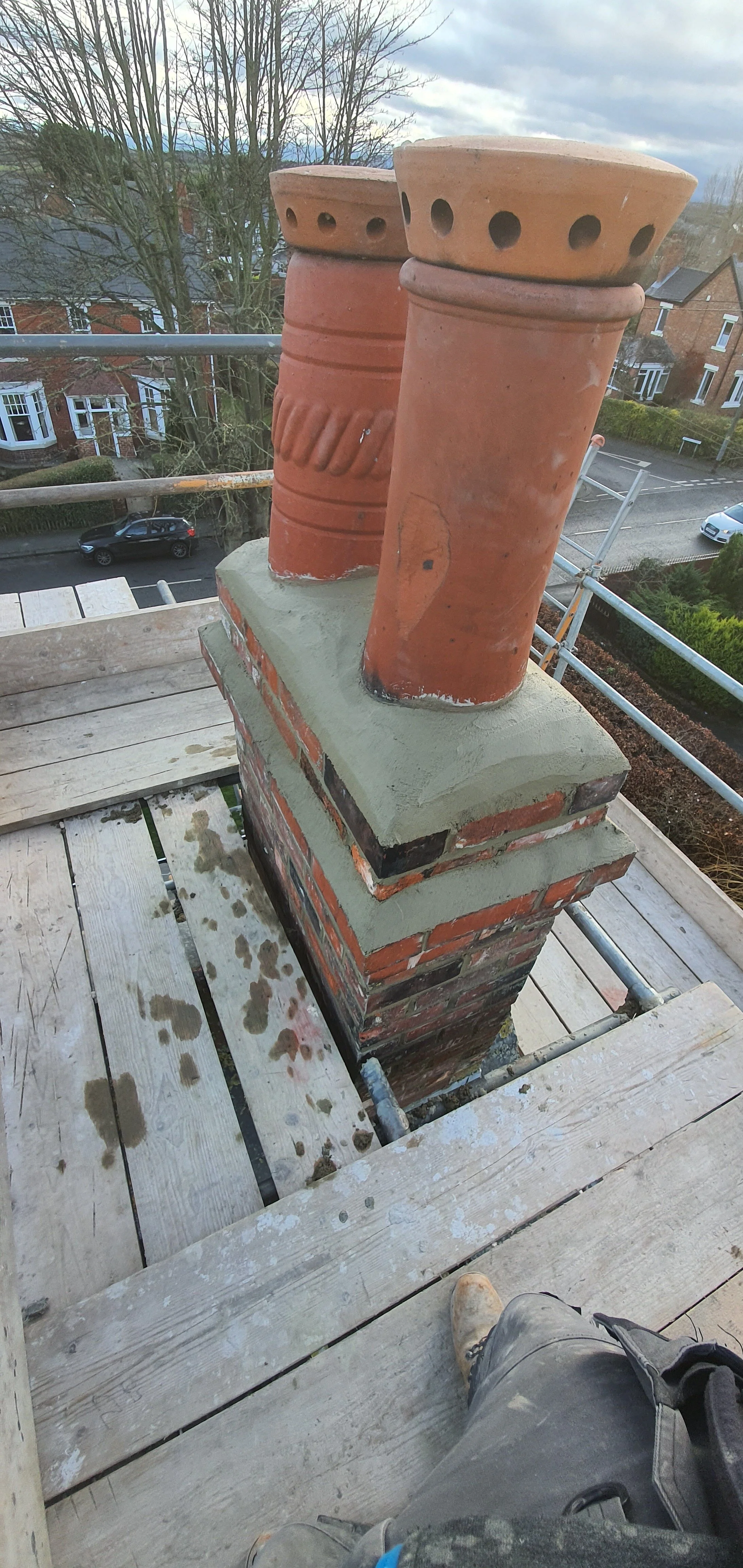 A chimney with two terracotta pots on top, situated on a brick fireplace base, seen from a wooden scaffolding platform on a residential street with houses and cars visible in the background.
