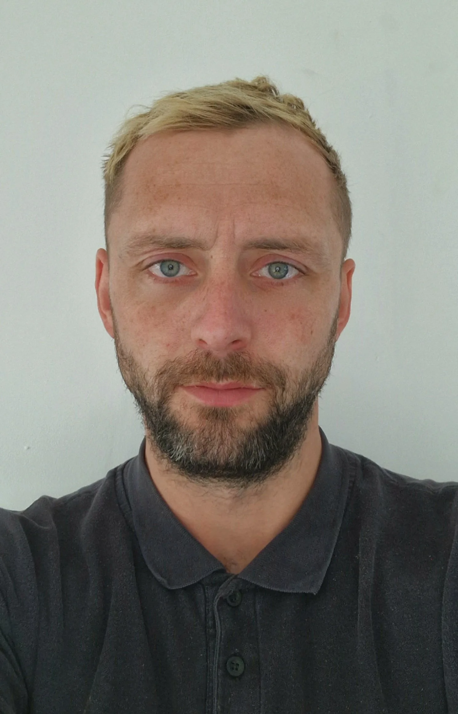 A man with short blond hair and a beard taking a selfie against a plain light-colored wall.