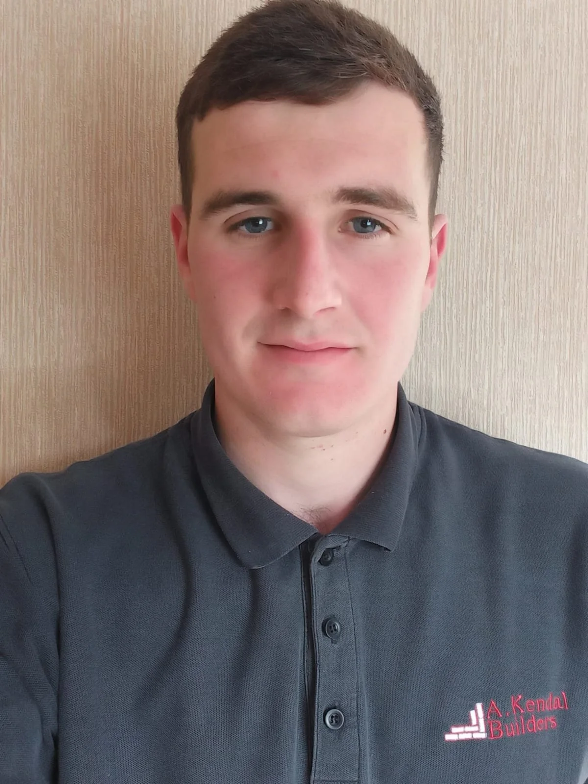 A young man with short brown hair, blue eyes, and fair skin, wearing a dark gray polo shirt with red and white logo embroidery that reads "A. Kendal Builders," standing in front of a wooden textured wall.