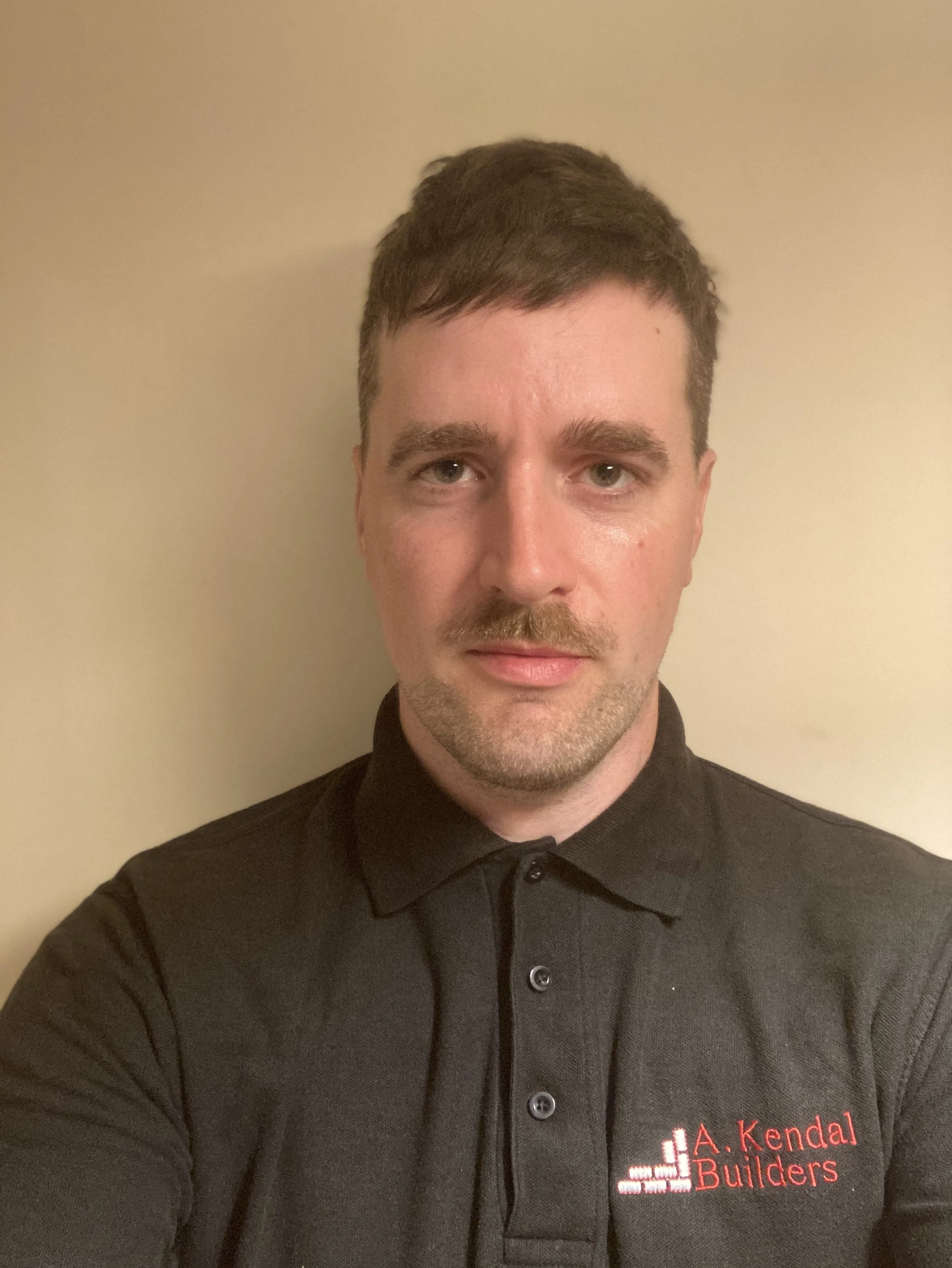 A man taking a selfie against a beige wall, wearing a black polo shirt with the logo of A. Kendal Builders.