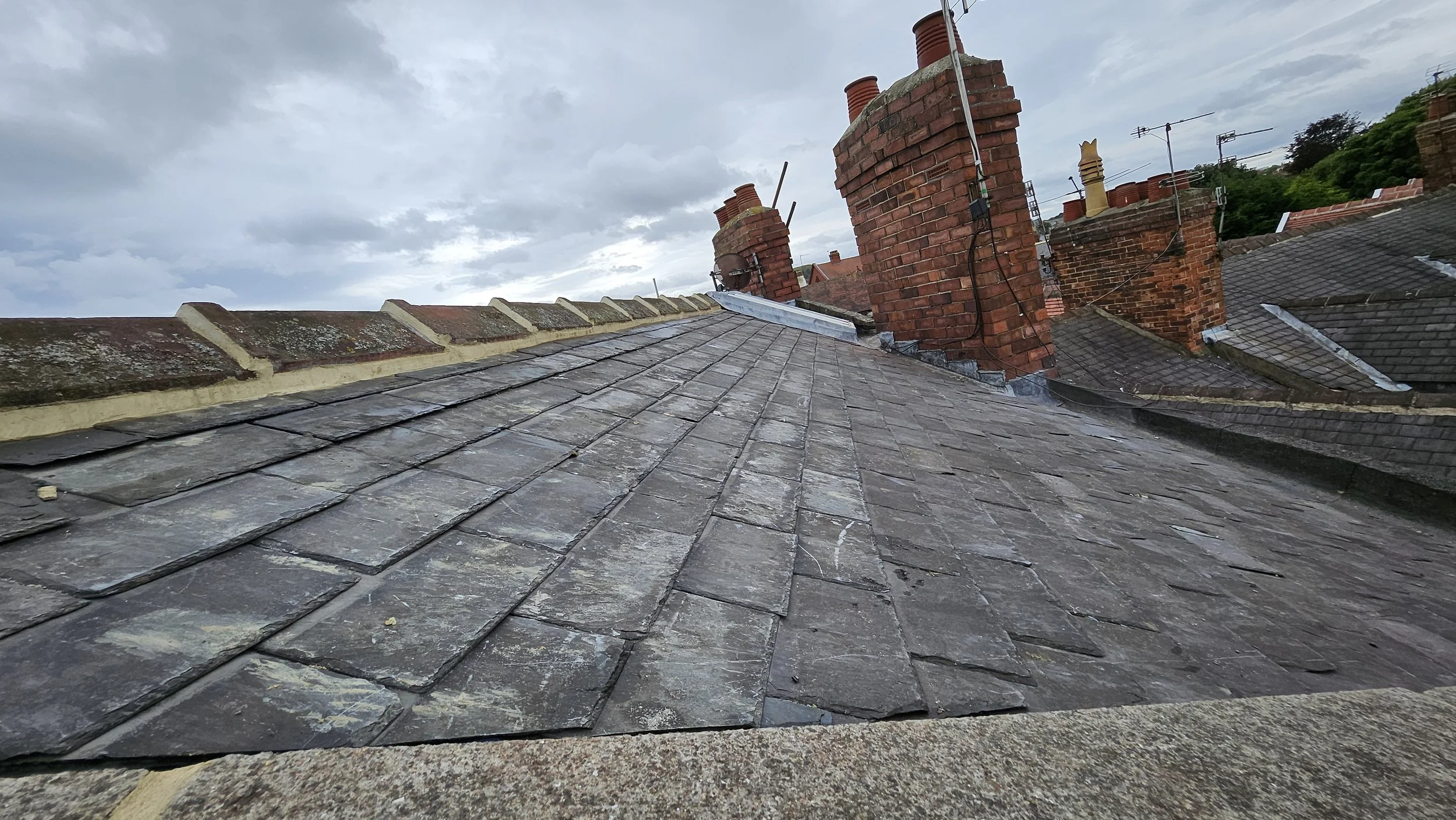View of slate roof tiles and multiple brick chimneys on a rooftop under cloudy sky.