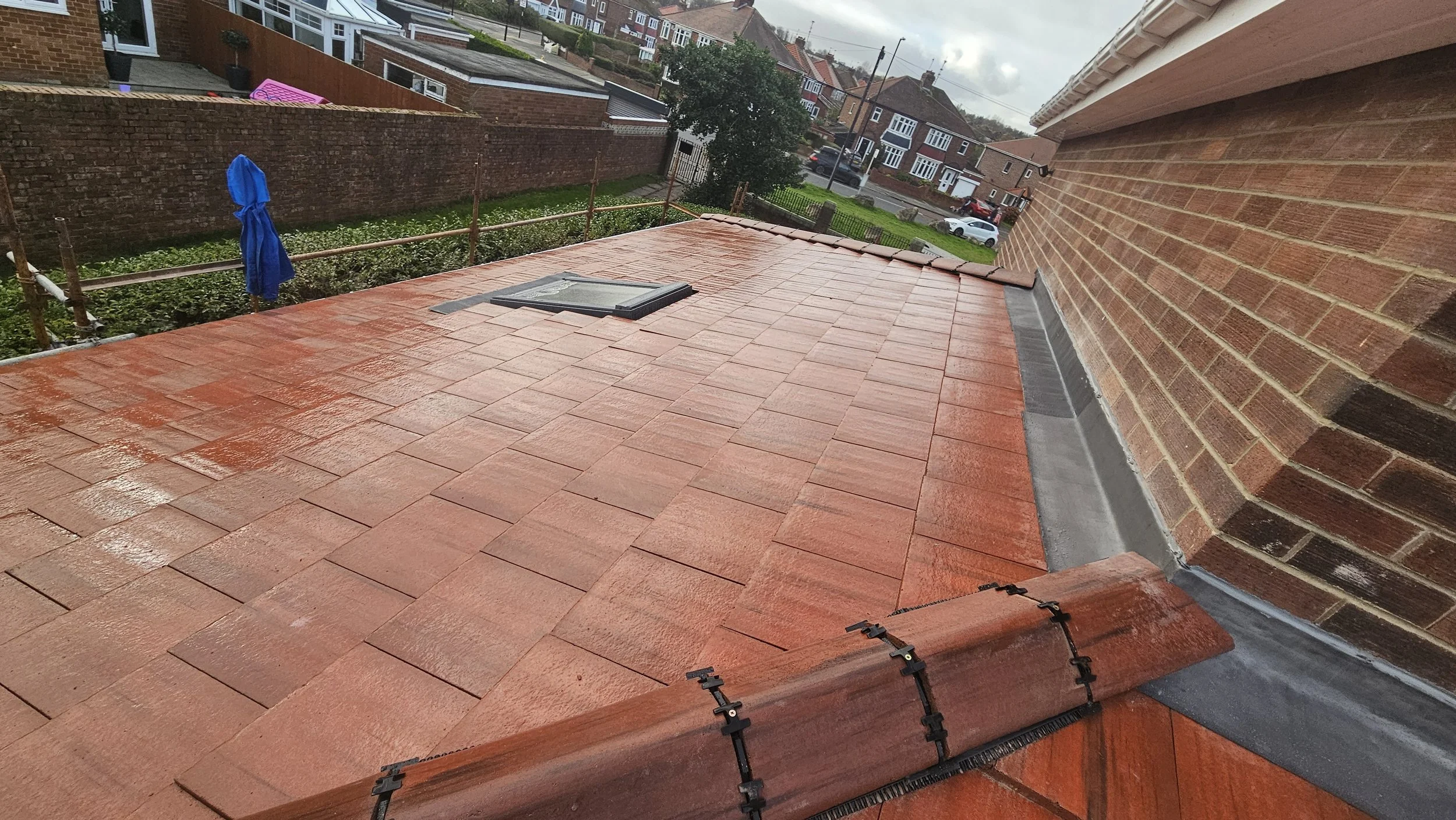 A newly installed red brick or tile roof on a building, featuring a roof window and a gutter along the edge, with residential houses and a street visible in the background on a cloudy day.