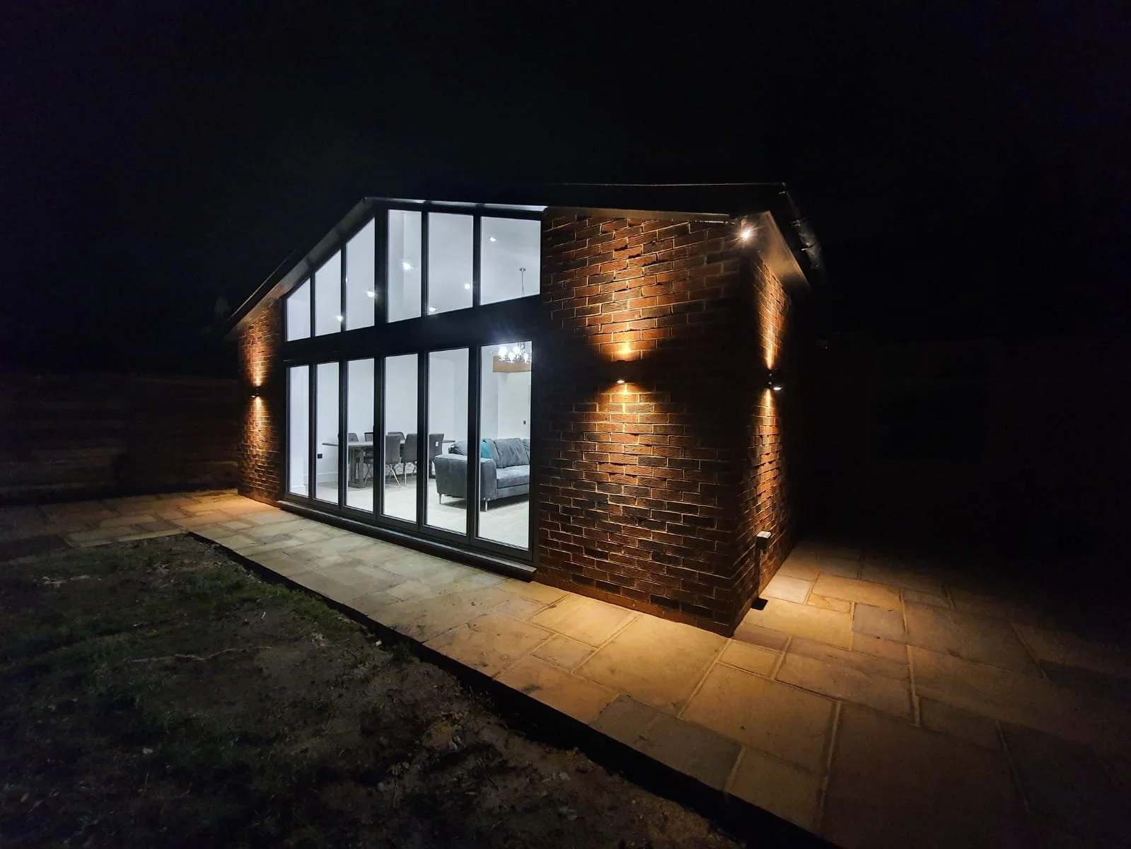 A modern brick and glass house at night with interior lighting visible through large windows, surrounded by a paved area and some grass.