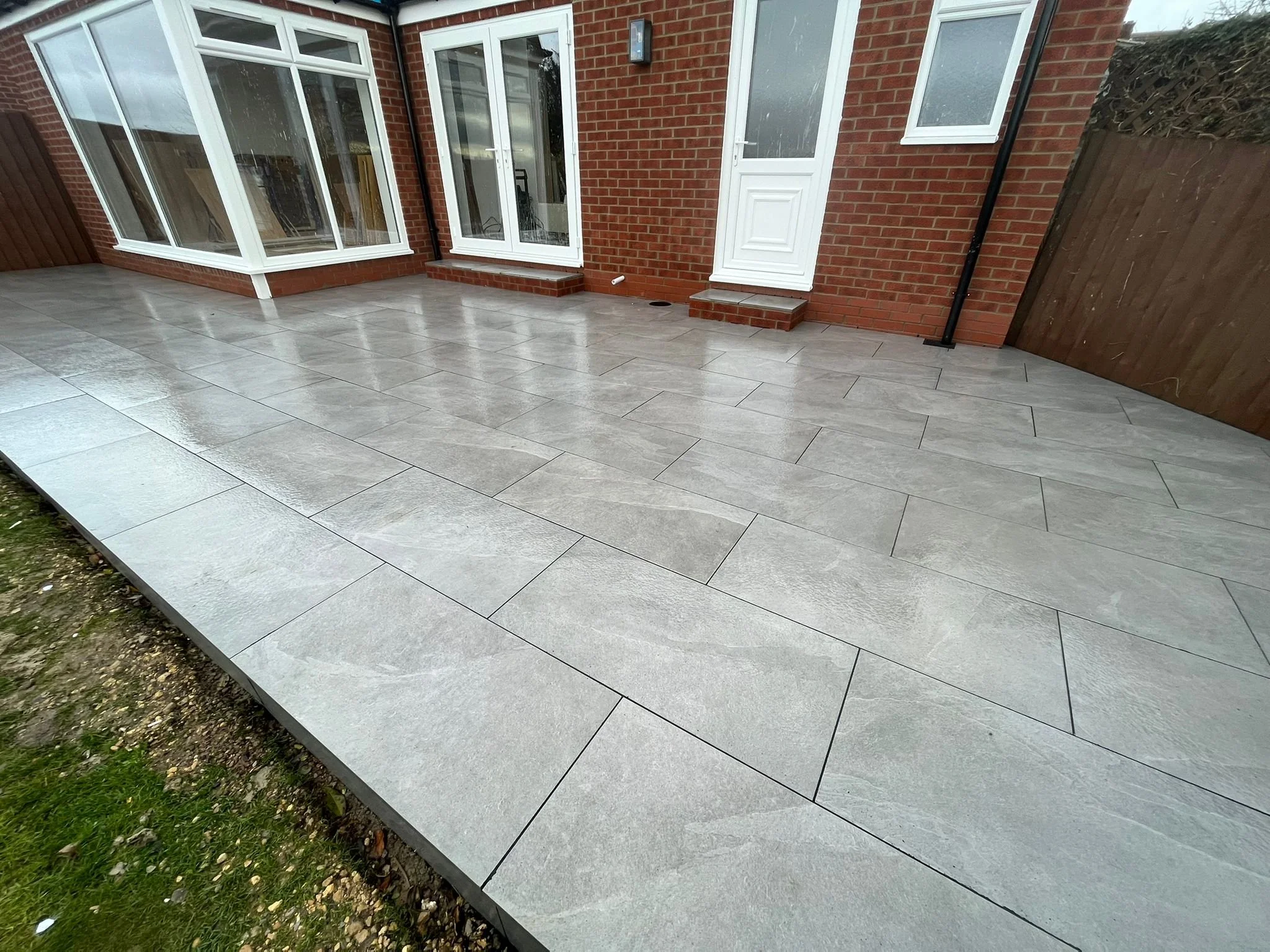 Newly installed light gray tiled patio outside brick house with white door and windows, wooden fence on right, and small section of grass and dirt at the edge.