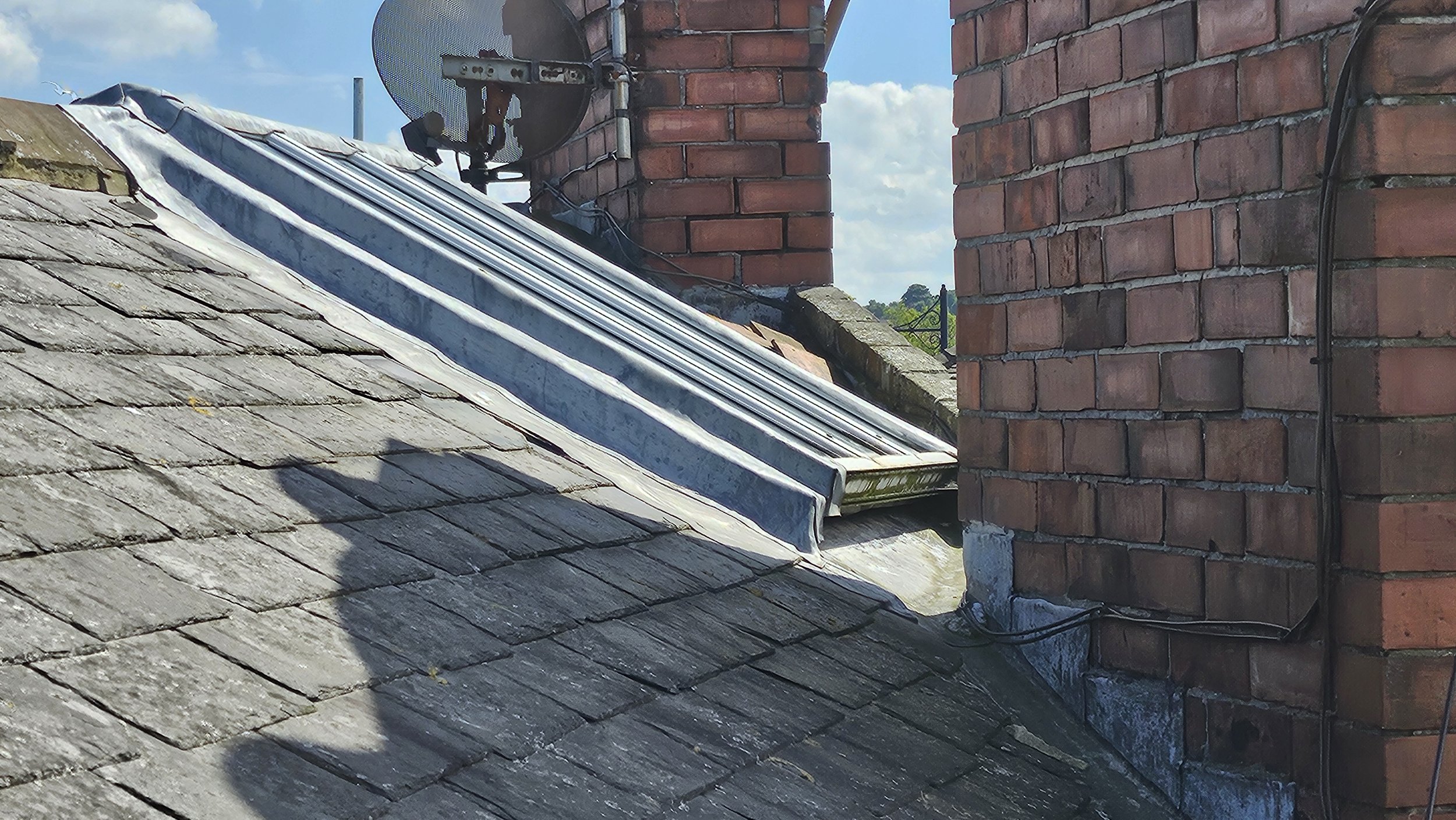 Close-up of a sloped tiled roof with a metal flashing installed along the roof ridge, and two red brick chimneys with electrical wiring attached. Satellite dish mounted near the top of one chimney. Blue sky with clouds in background.
