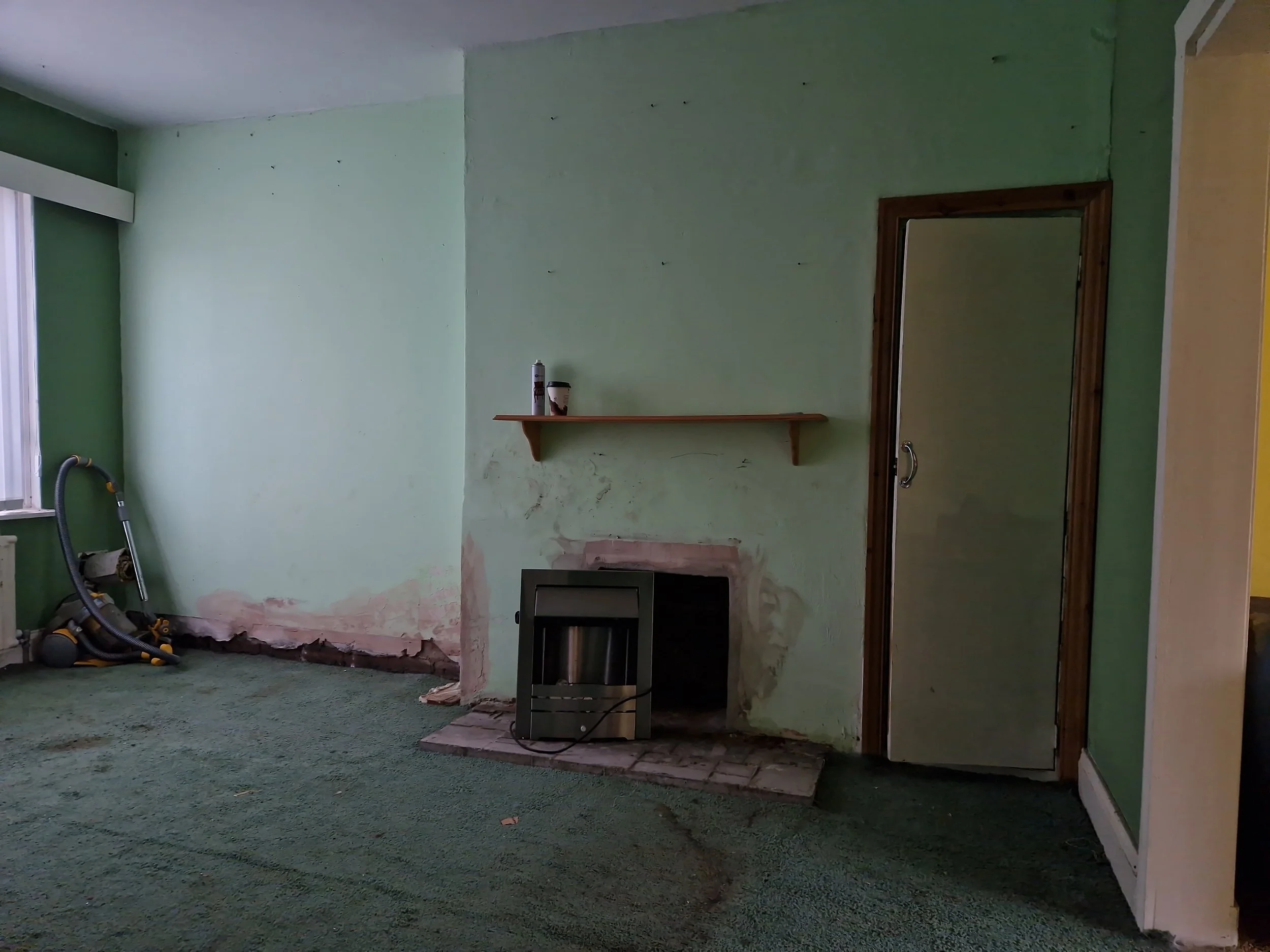 Living room with green walls, a green carpet, a small fireplace, a shelf with an item on it, a mirror, and a vacuum cleaner near the window.