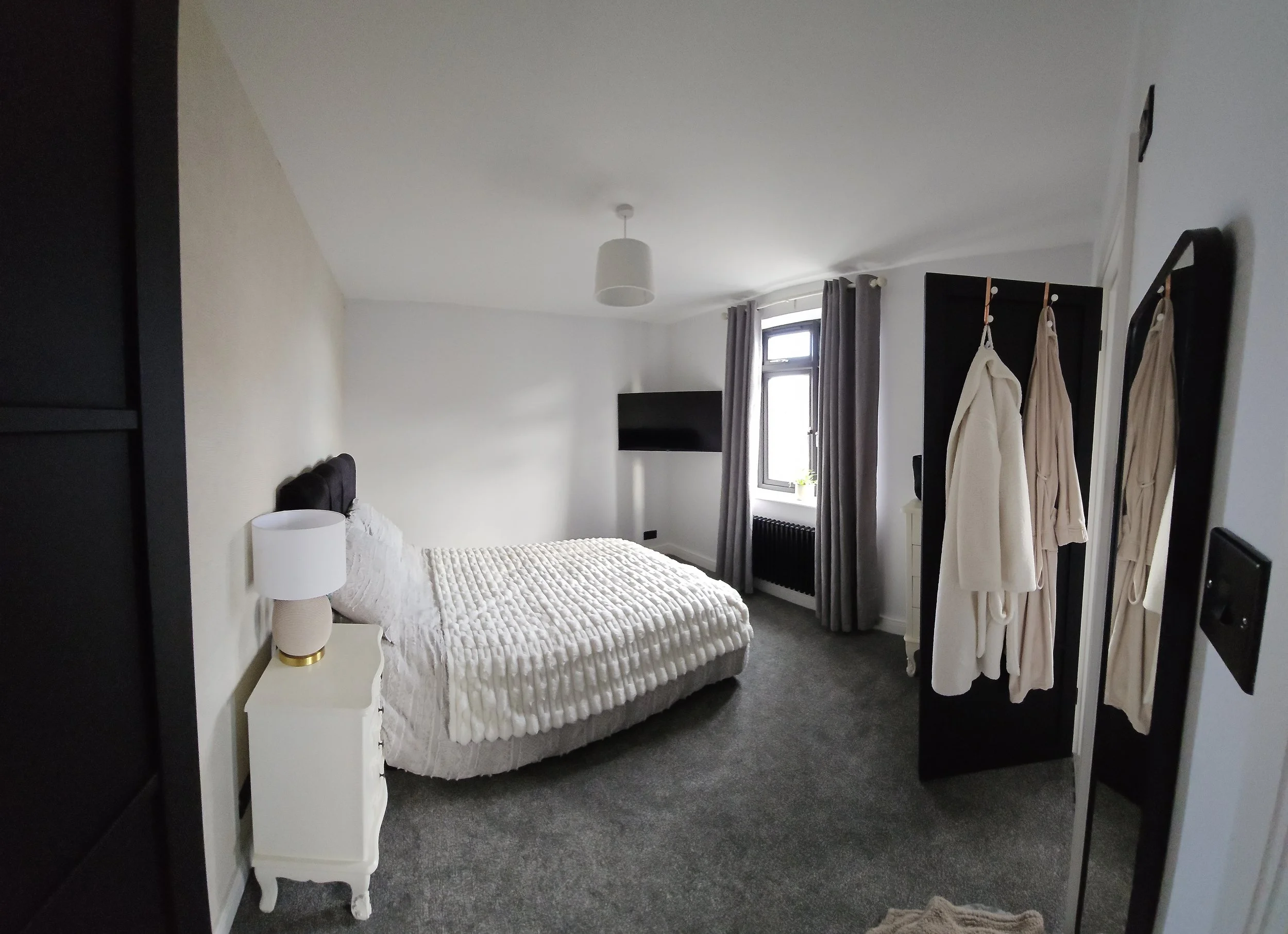 A bedroom with a bed, nightstand, lamp, window with curtains, mounted TV, and a black wardrobe with robes hanging on it.