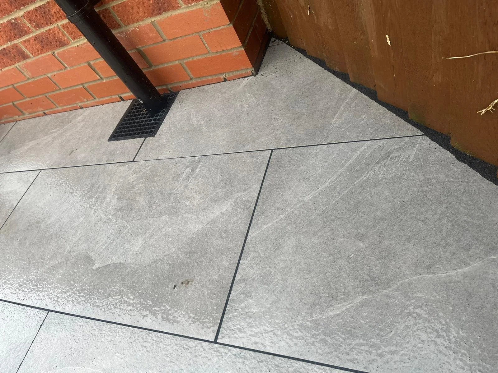 Close-up of wet grey paving stones on a sidewalk next to a brick wall and wooden fence, with a black drain grate and a black pipe.