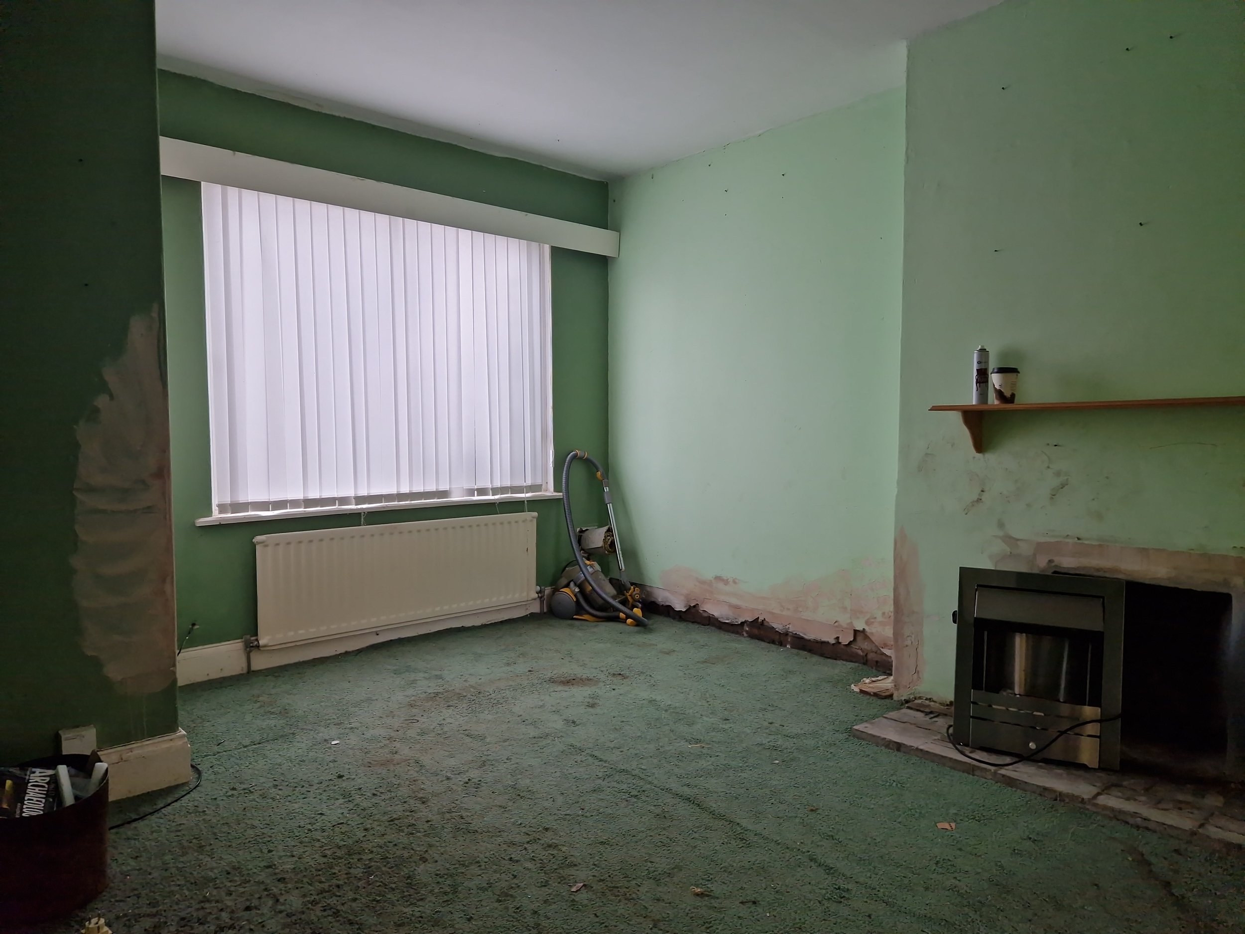 Empty room with green walls, a radiator under a window with vertical blinds, a fireplace on the right, and a vacuum cleaner leaning against the wall.