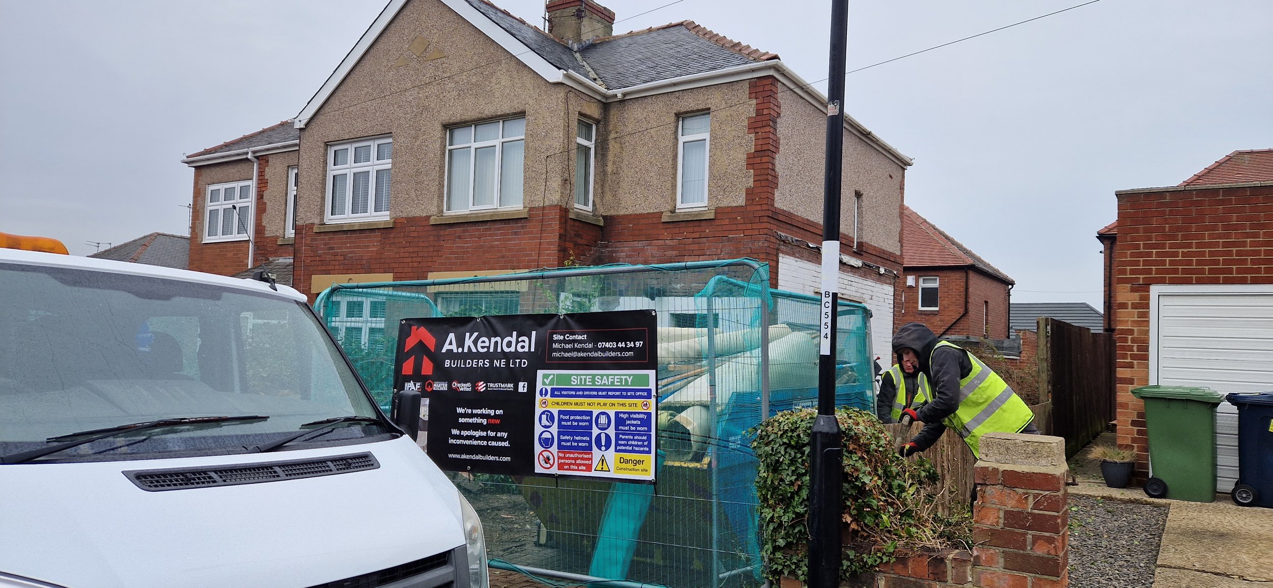 Construction workers in yellow safety vests working on a small brick wall outside a house under renovation. There is a sign on the fence indicating site safety and contact details of A. Kendal Builders Ltd. A white van is parked nearby, and in the ba