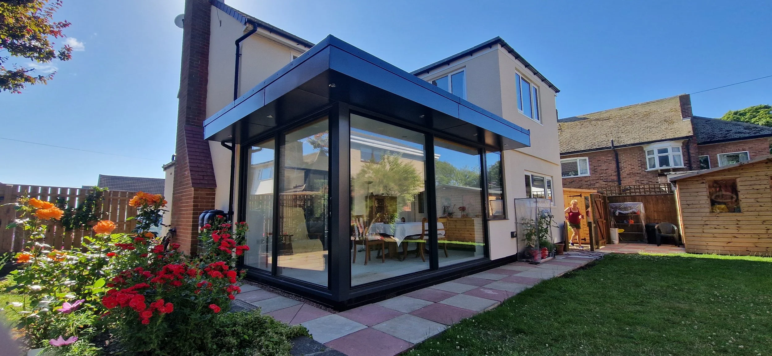 A modern extension with large glass windows attached to a house, surrounded by a garden with colorful flowers and a lawn, with a woman walking in the background.