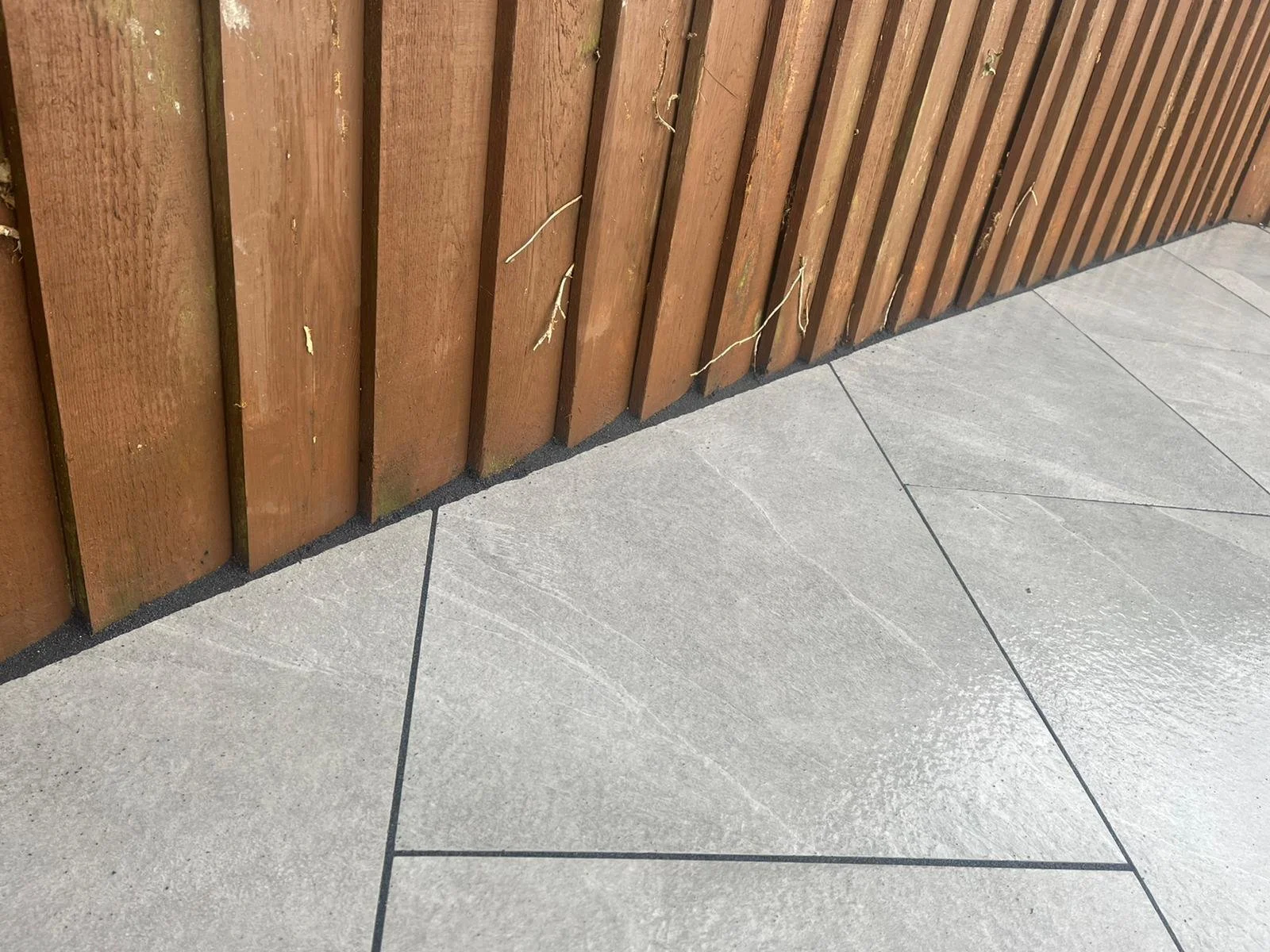 Close-up of a wooden fence with some small twigs and leaves, adjacent to a tiled concrete surface.