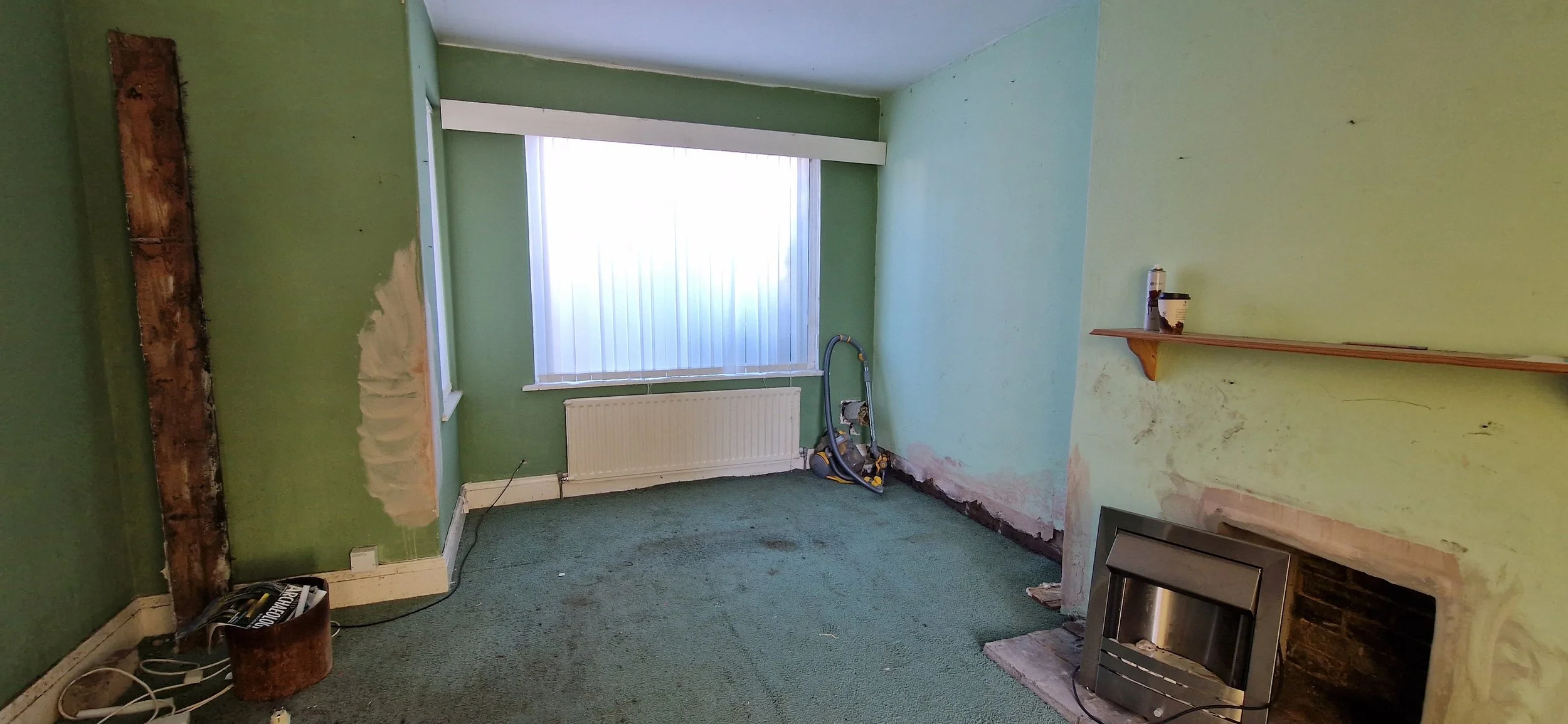 Empty room with green walls and green carpet, partially renovated with exposed brick and wall patches, a window with blinds, a small shelf on the wall, an old fireplace, and a vacuum cleaner on the floor.