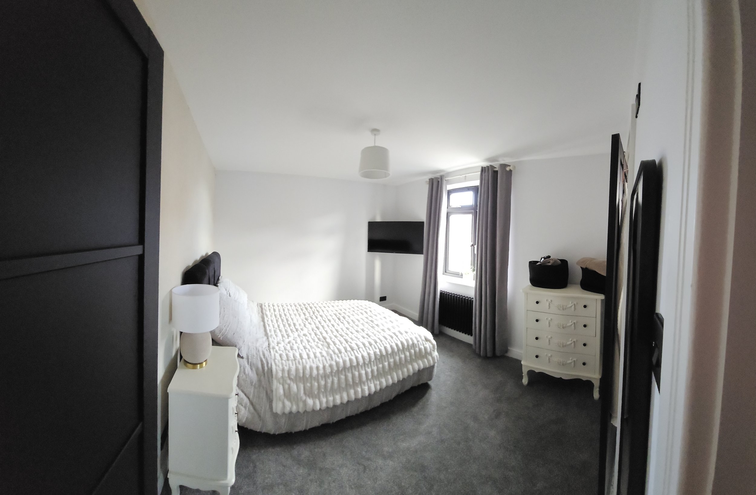 A bedroom with a bed, nightstand with lamp, dresser, wall-mounted TV, window with curtains, and gray carpet.