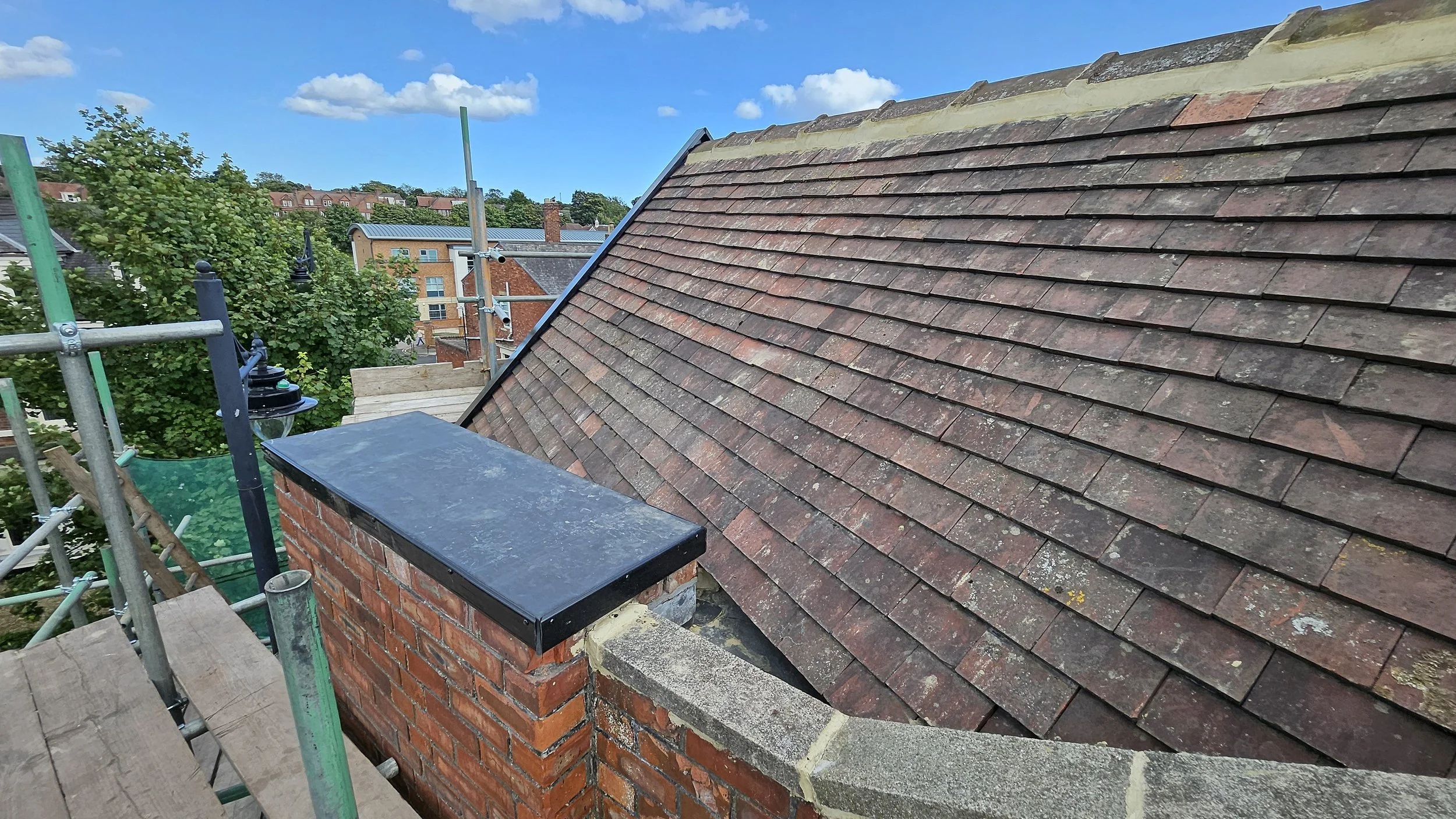 Roof with red and brown shingles, brick chimney with black cap, scaffolding and green netting nearby, and trees and buildings in the background under a partly cloudy blue sky.
