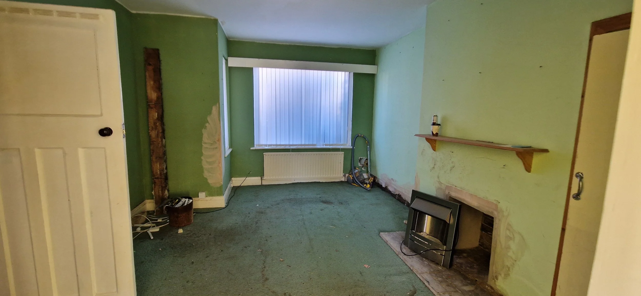 An empty living room with green walls, a window with closed vertical blinds, and a green carpet. The room has a partially removed fireplace with visible brick and a TV placed in front of it. There is a wooden shelf on the wall, an old vent, and a few