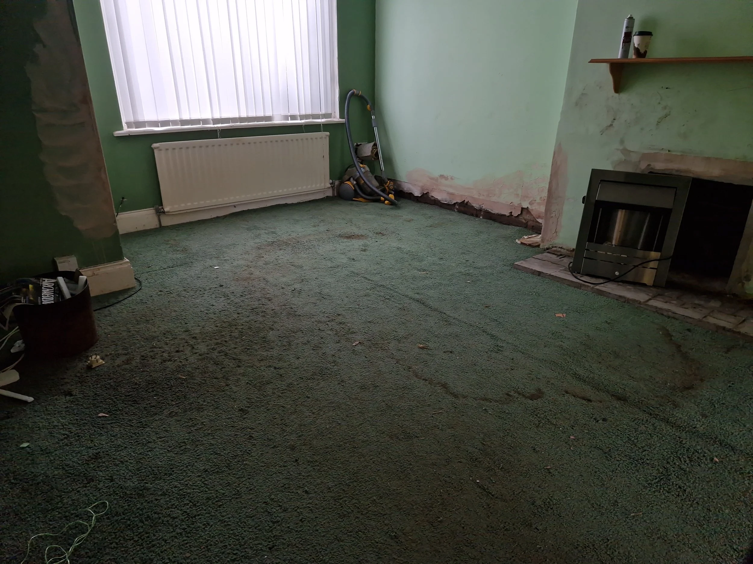 A vacant room with green walls and carpet flooring, partially removed near a fireplace, with a vacuum cleaner in the corner, a window with vertical blinds, and cluttered items on the floor.