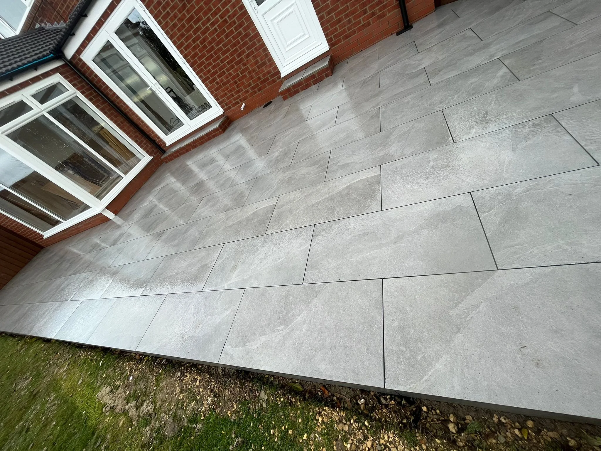 Newly paved patio with large beige tiles in front of a brick house with large windows and a white door.