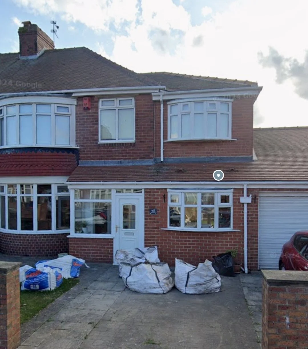 Front view of a two-story brick house with bay windows, a driveway with three large bags of yard waste, and a closed garage door on the right.
